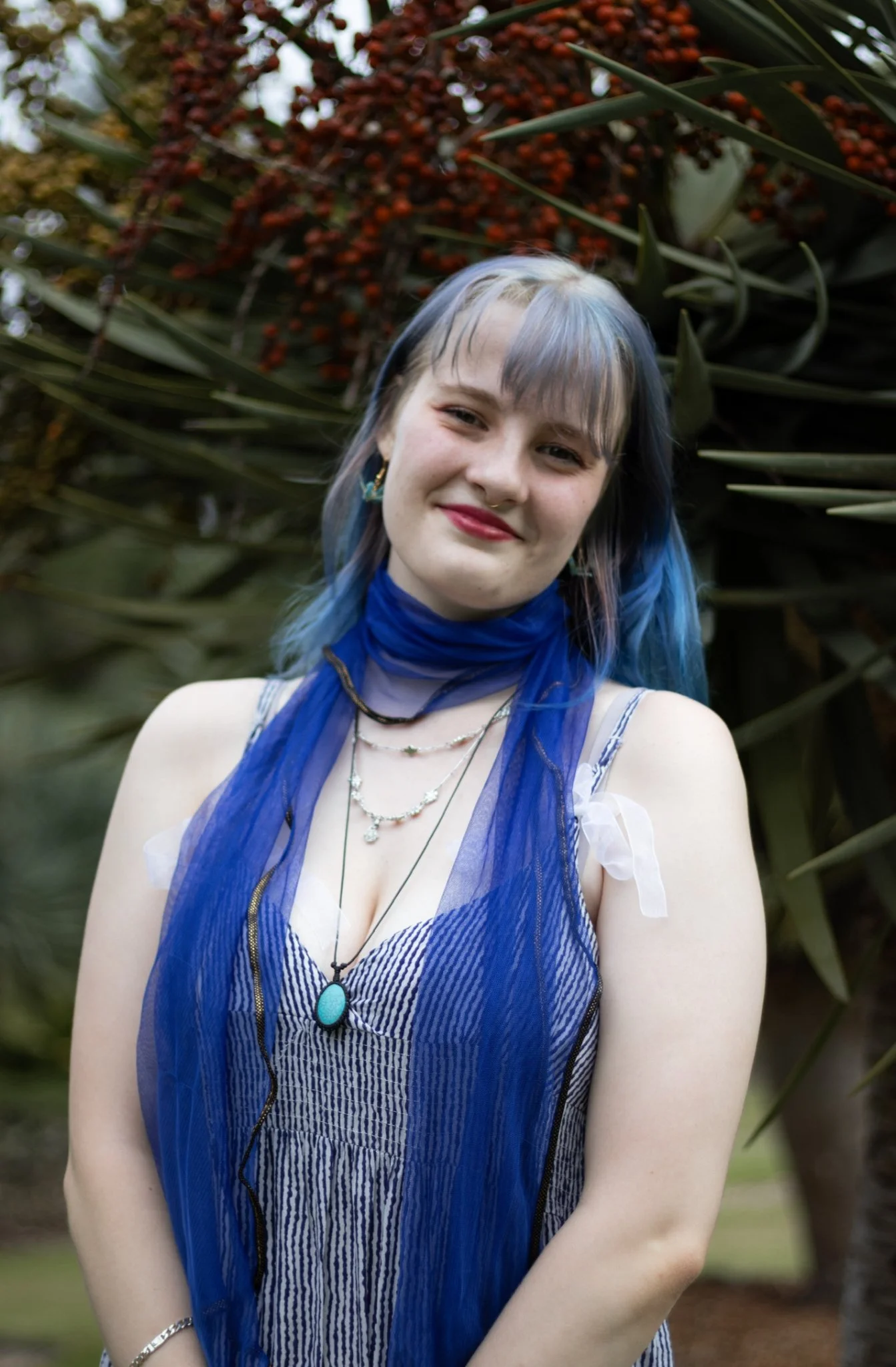 A woman with blue hair, wearing a striped dress, layered necklaces, and a blue sheer scarf, stands outdoors in front of green plants and red flowers, smiling at the camera.