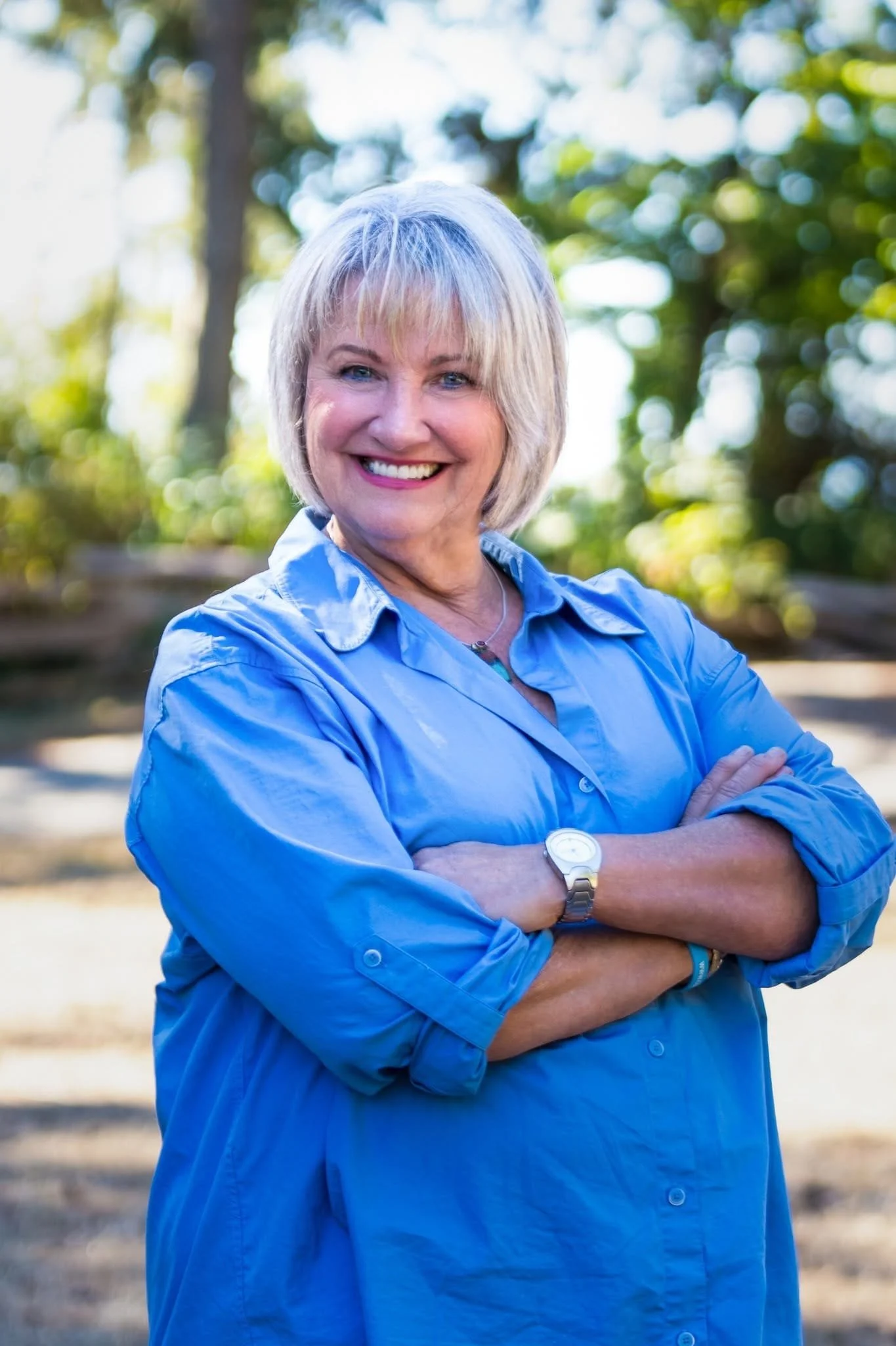 A smiling older woman with short blonde hair wearing a light blue shirt, standing outdoors with blurred green trees and sunlight in the background.