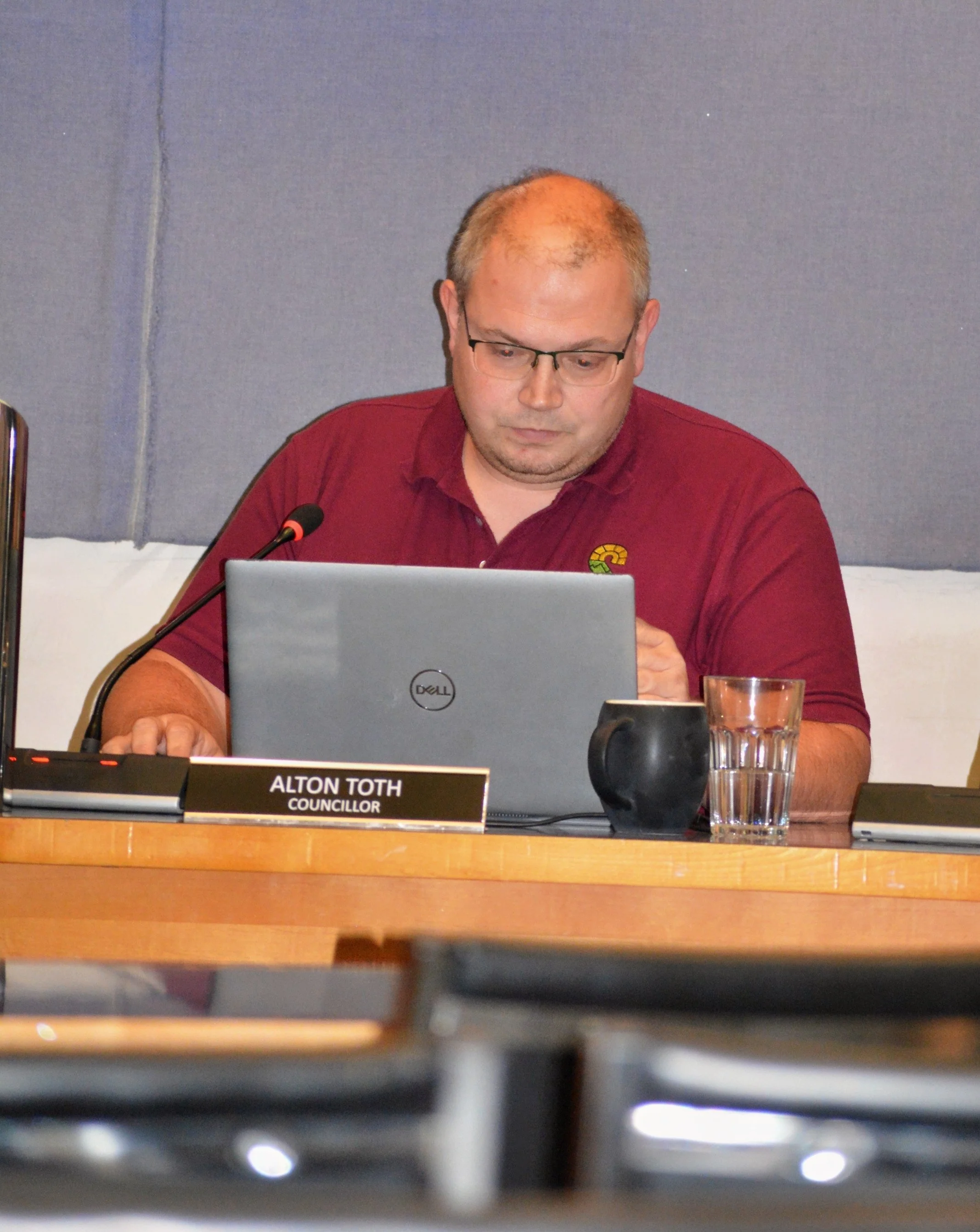A man with glasses and a red shirt sitting at a table, looking at a laptop, with a nameplate that reads 'Alton Toth, Councillor', a microphone, a glass of water, and a smartphone in front of him.