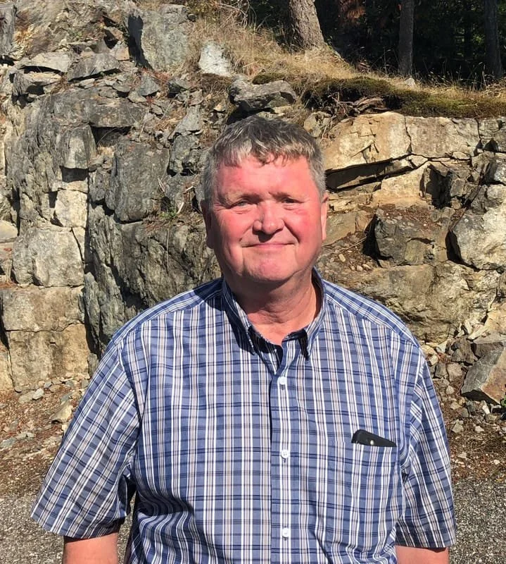 A man with short gray hair wearing a blue and white plaid button-up shirt standing outdoors in front of a rocky hillside.
