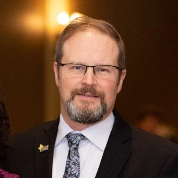 A middle-aged man with glasses, a beard, and light brown hair, dressed in a suit and tie, standing indoors at an event.