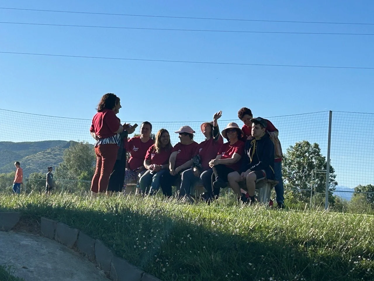 Group of people sitting on a bench outdoors, some wearing hats, with a woman standing and speaking to them, in a grassy area with a sports net and hills in the background on a sunny day.
