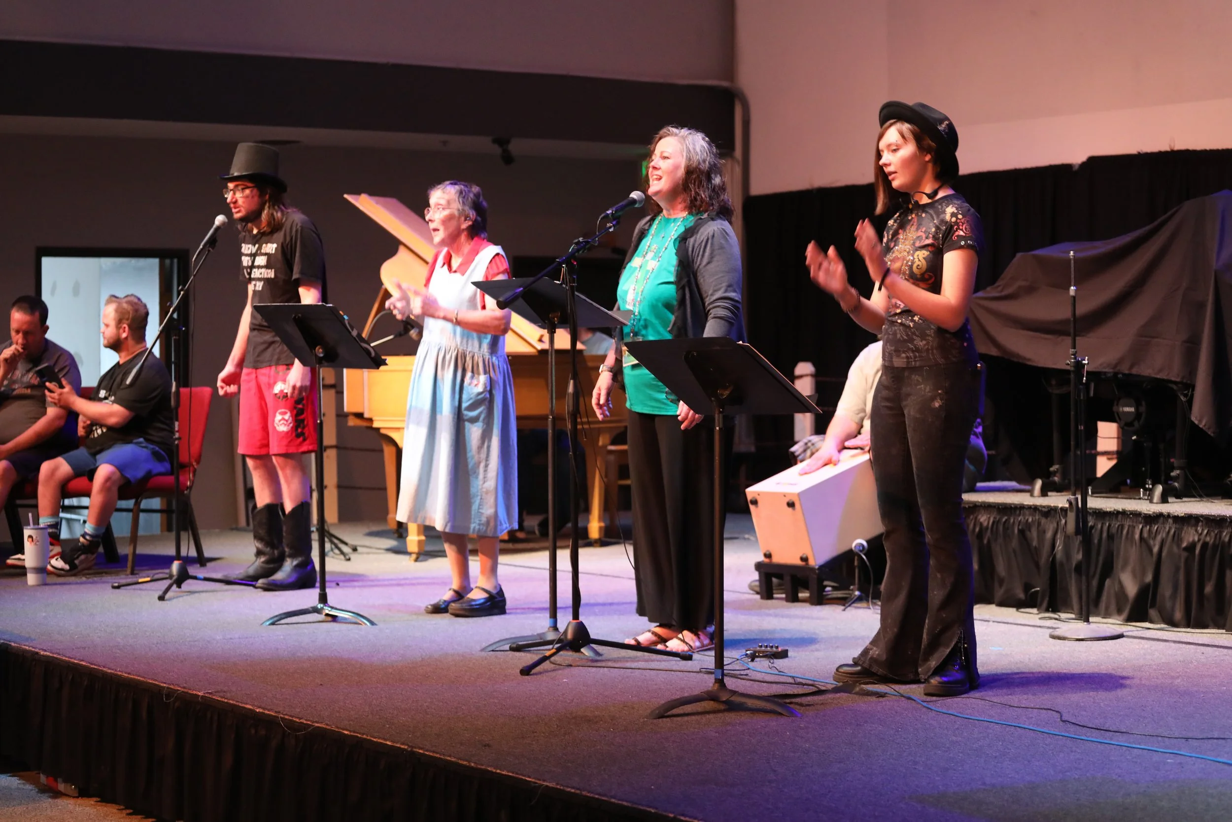 Four people on stage at a performance, three women and one man, with musical stands and a piano in the background. One woman is speaking or singing into a microphone, the others are standing or clapping.