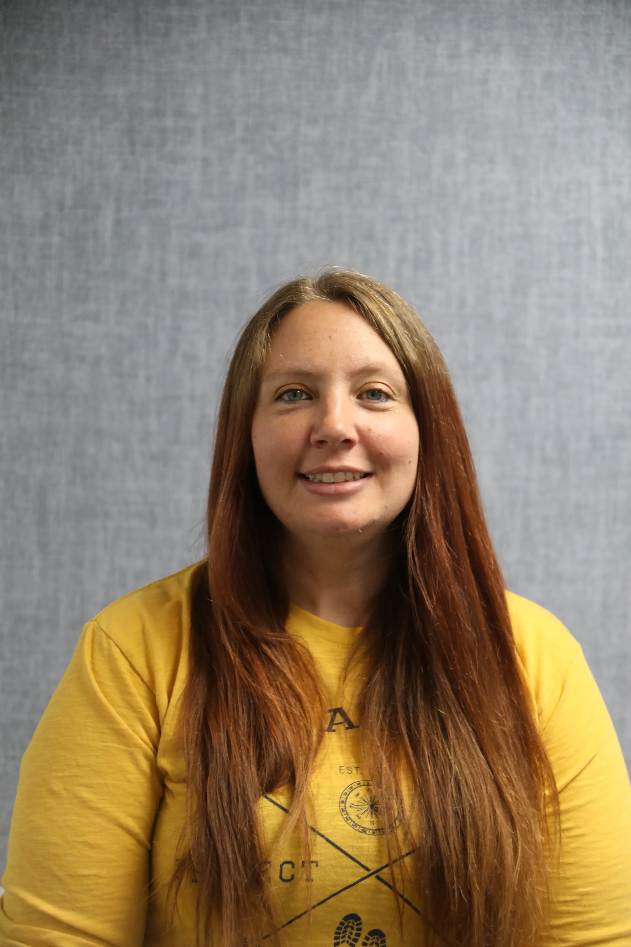 A woman with long red hair wearing a yellow shirt, smiling, in front of a grey background.