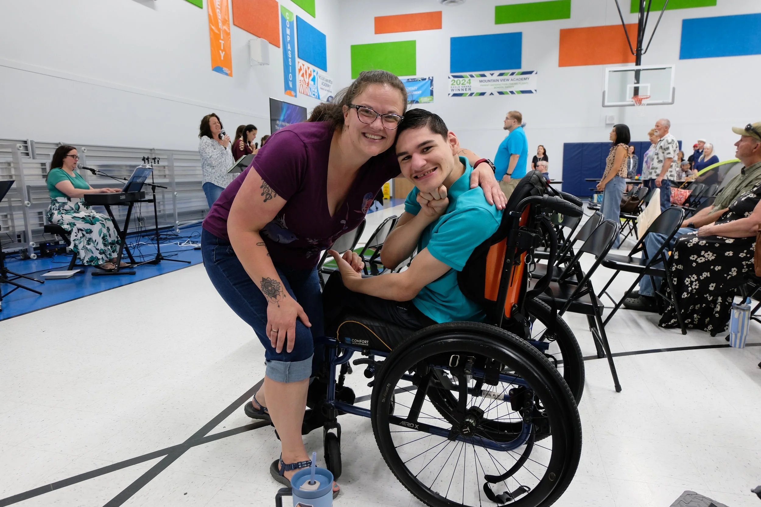 A woman with glasses and a tattoo smiling and hugging a young man in a wheelchair at a gymnasium or community center, with people in the background participating in activities or waiting.