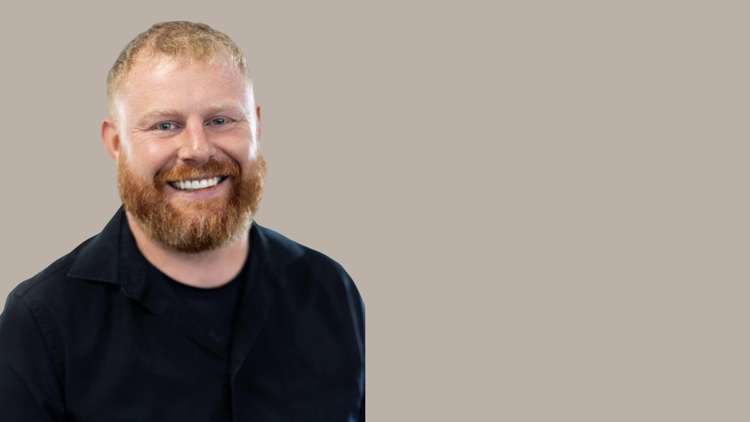 A smiling man with red hair and beard wearing a black shirt, with a neutral background.