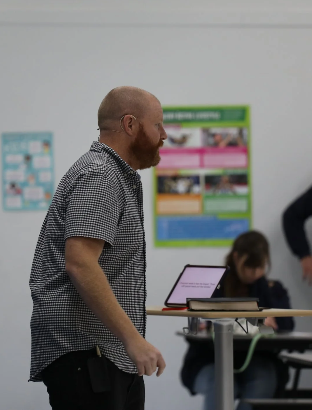 A man with a beard and checked shirt standing near a tablet on a stand in a classroom with posters on the wall.