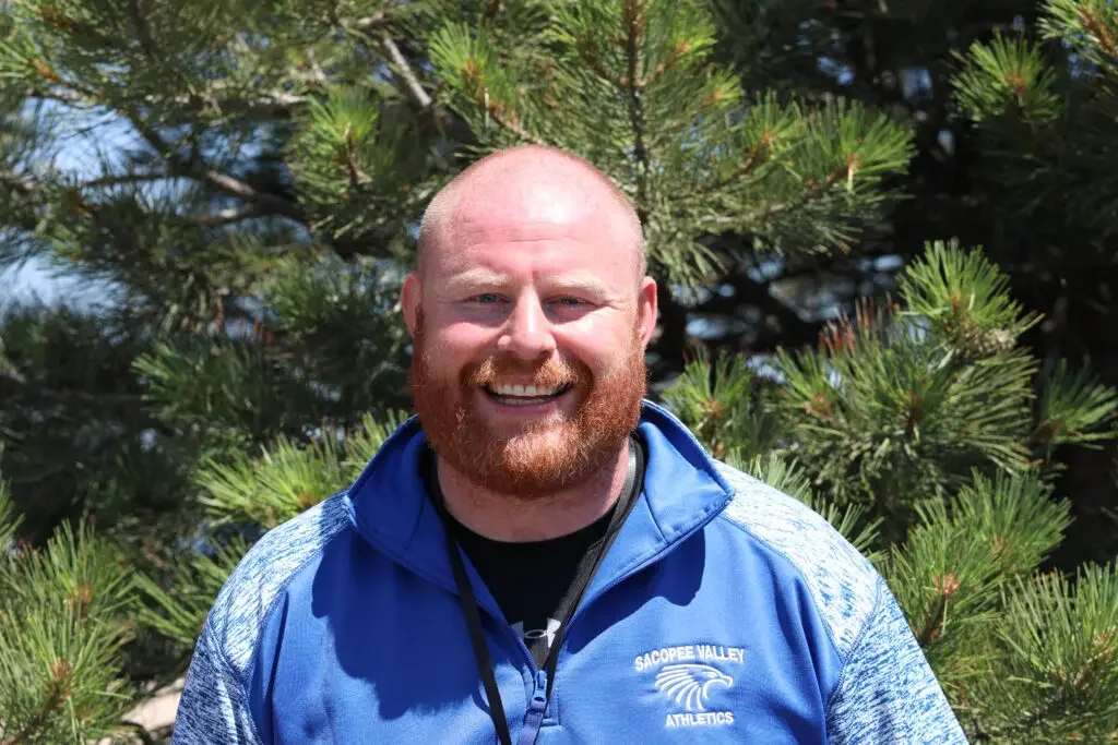 A smiling man with a red beard and shaved head standing outdoors in front of pine trees wearing a blue athletic jacket with 'Sacopee Valley Athletics' logo.
