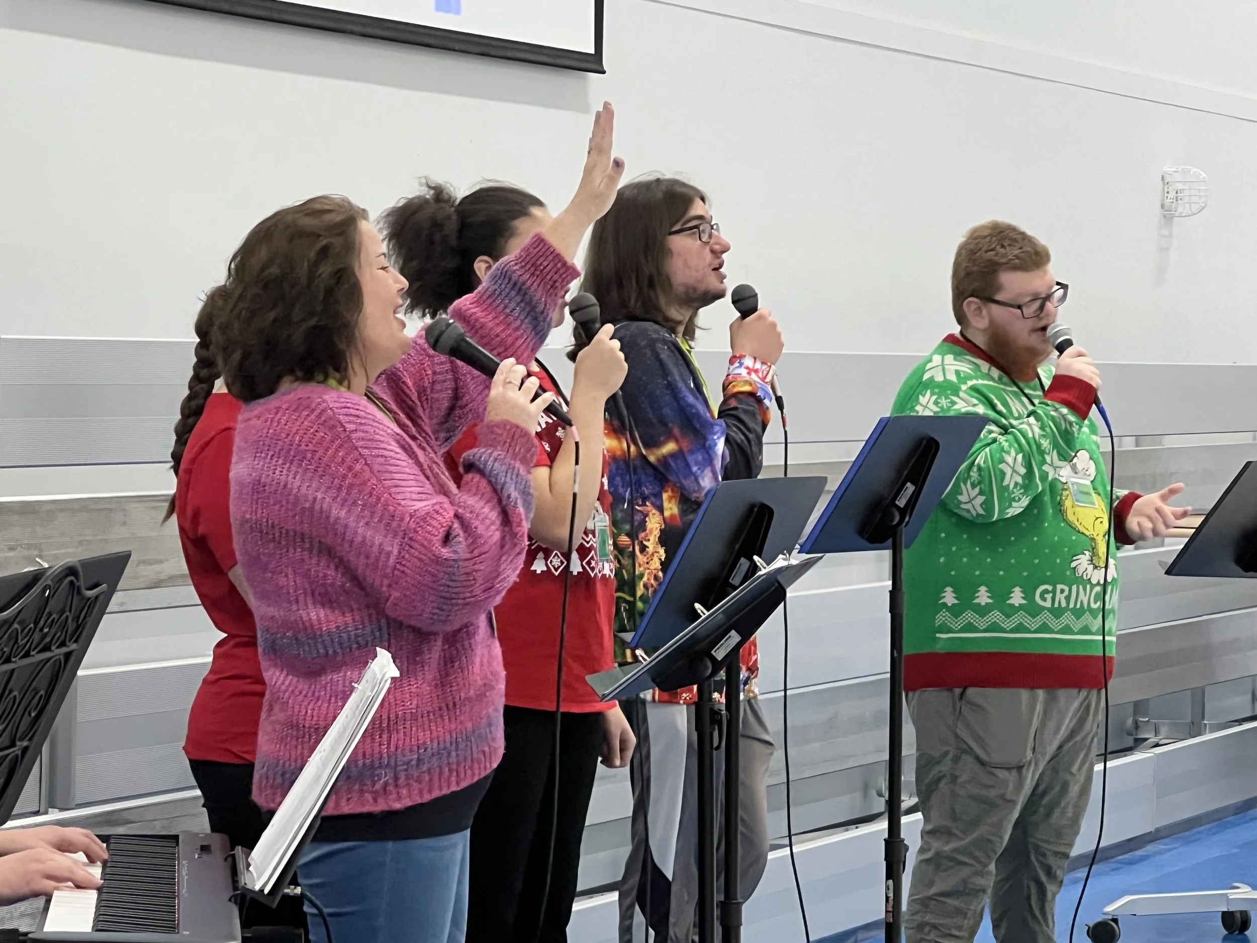 Group of four people singing into microphones, standing at music stands with sheet music, in a room with gray walls.
