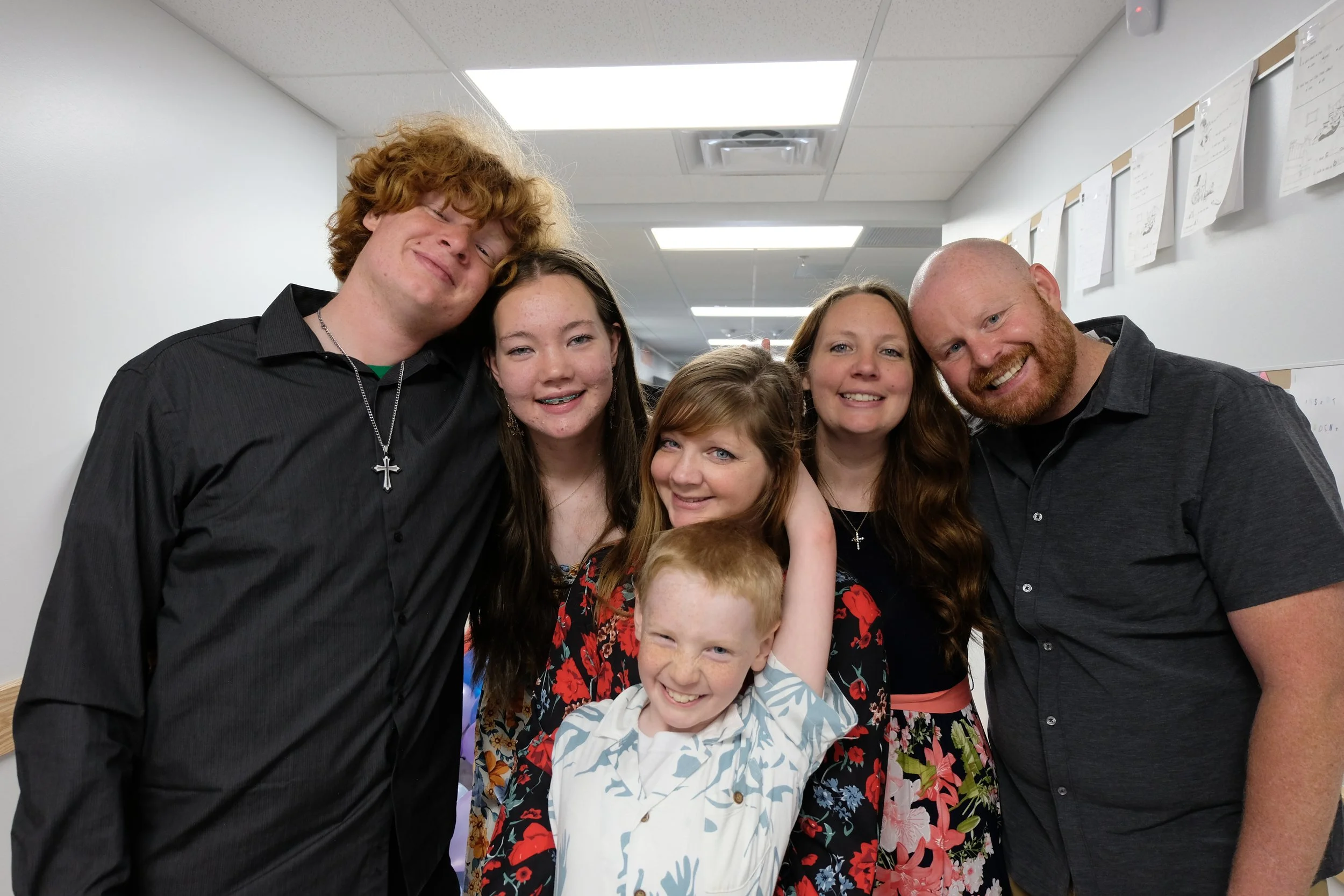 A group of six people smiling and posing closely together in an indoor setting. The group includes three adults and three children, with some leaning on each other. The background shows a white wall with papers or posters attached to it and ceiling lights.