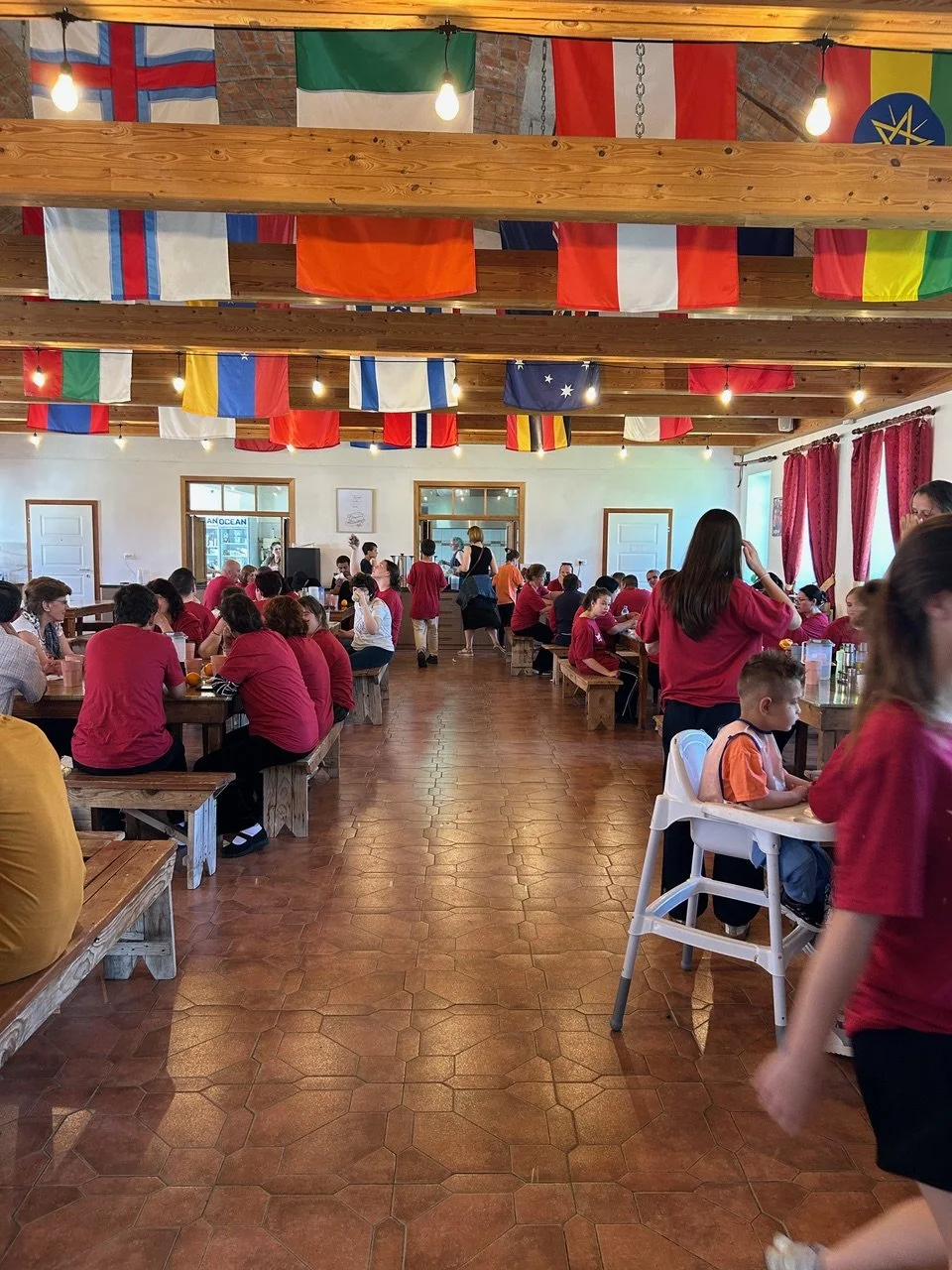 A busy dining hall filled with people sitting at wooden tables, some children and adults, under hanging international flags and string lights.