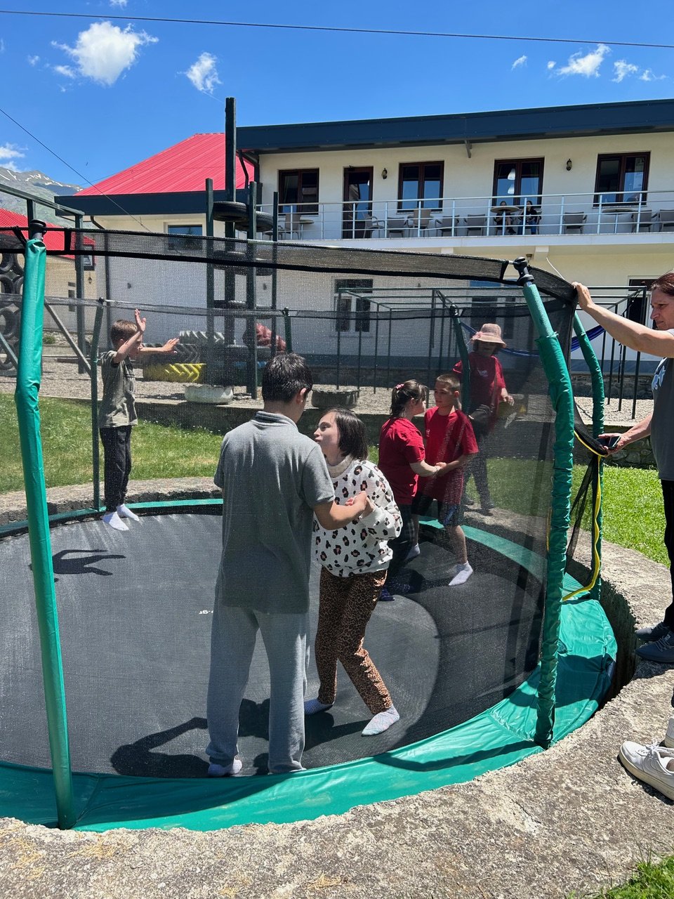 Children and adults playing on a trampoline outdoors on a sunny day with a modern house in the background.
