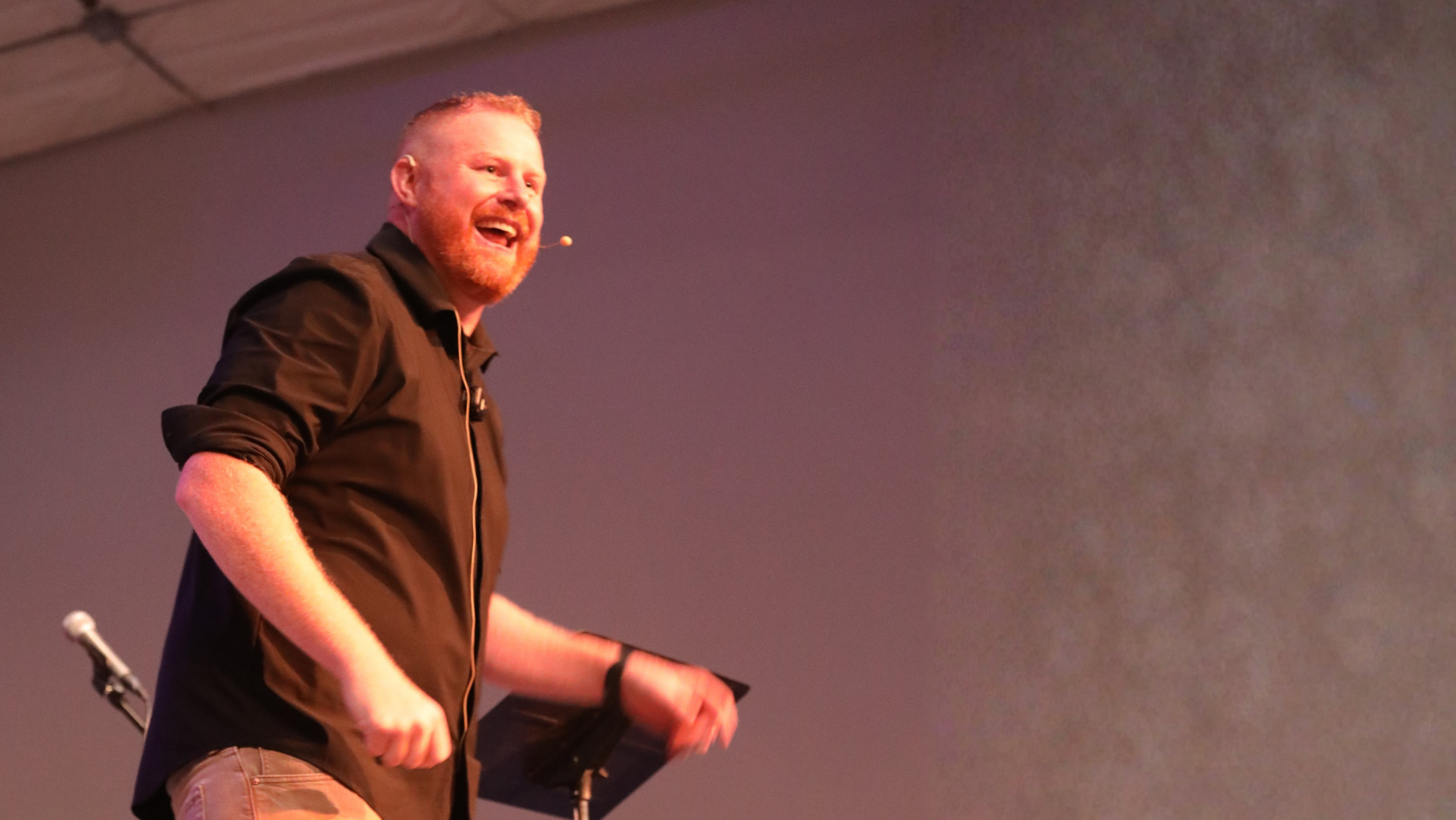 A man with red hair and a beard, wearing a black shirt, standing on stage, smiling and speaking into a microphone, with a purple background.