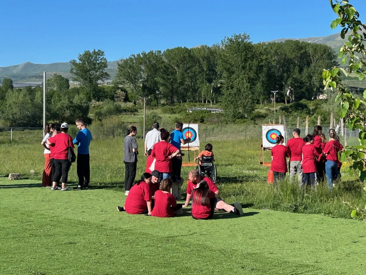 A group of people gathered outdoors in bright sunlight, some sitting on grass and others standing near archery targets, engaging in or observing an archery activity in a lush green field with trees and hills in the background.