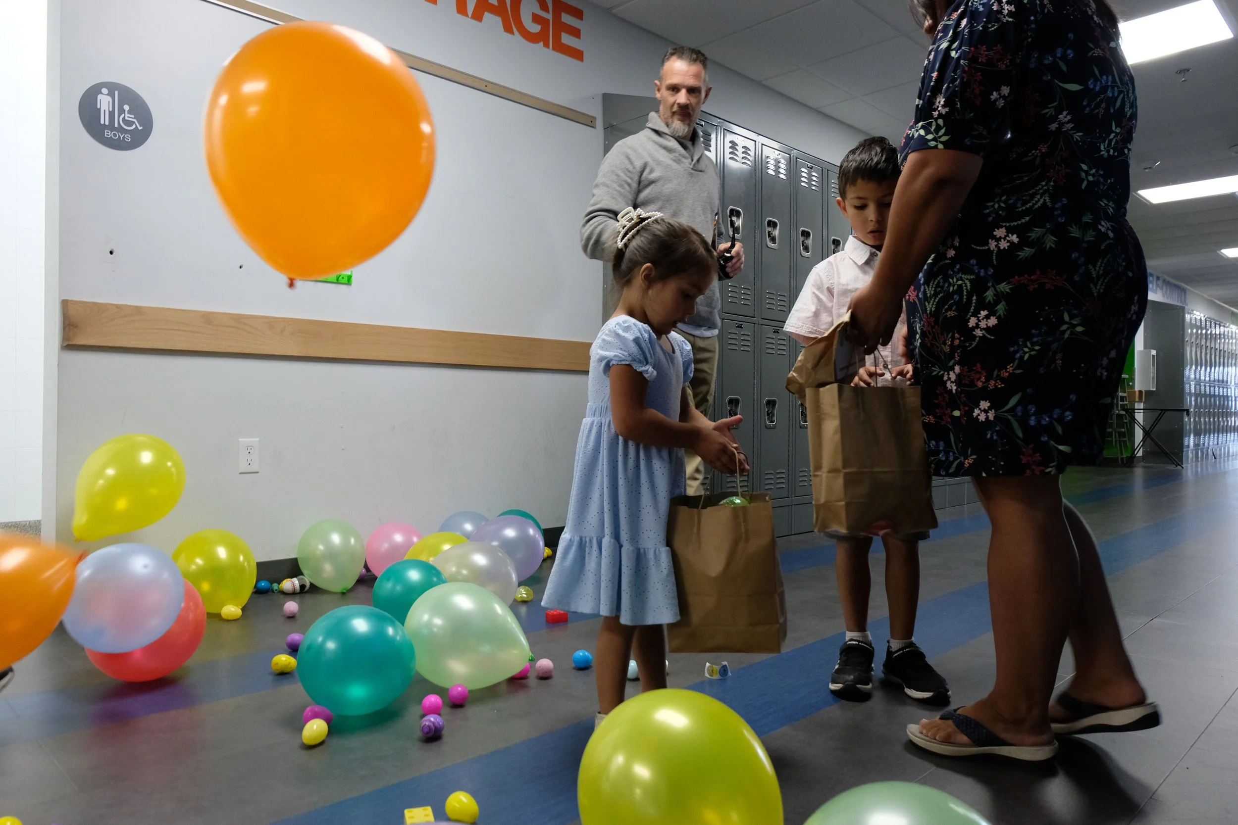 Children and adults at a school event with colorful balloons, some on the floor and hanging, in a hallway near lockers.