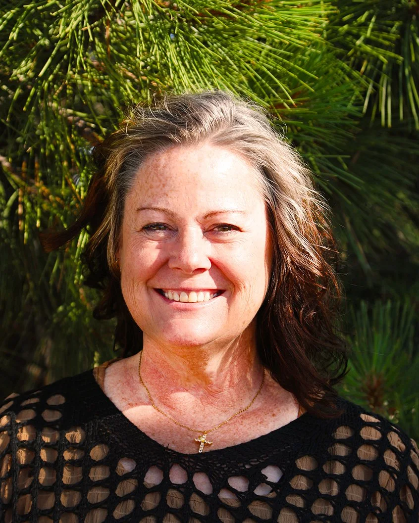 Smiling woman with light skin and shoulder-length wavy hair standing outdoors in front of green foliage, wearing a black crochet top and a gold cross necklace.