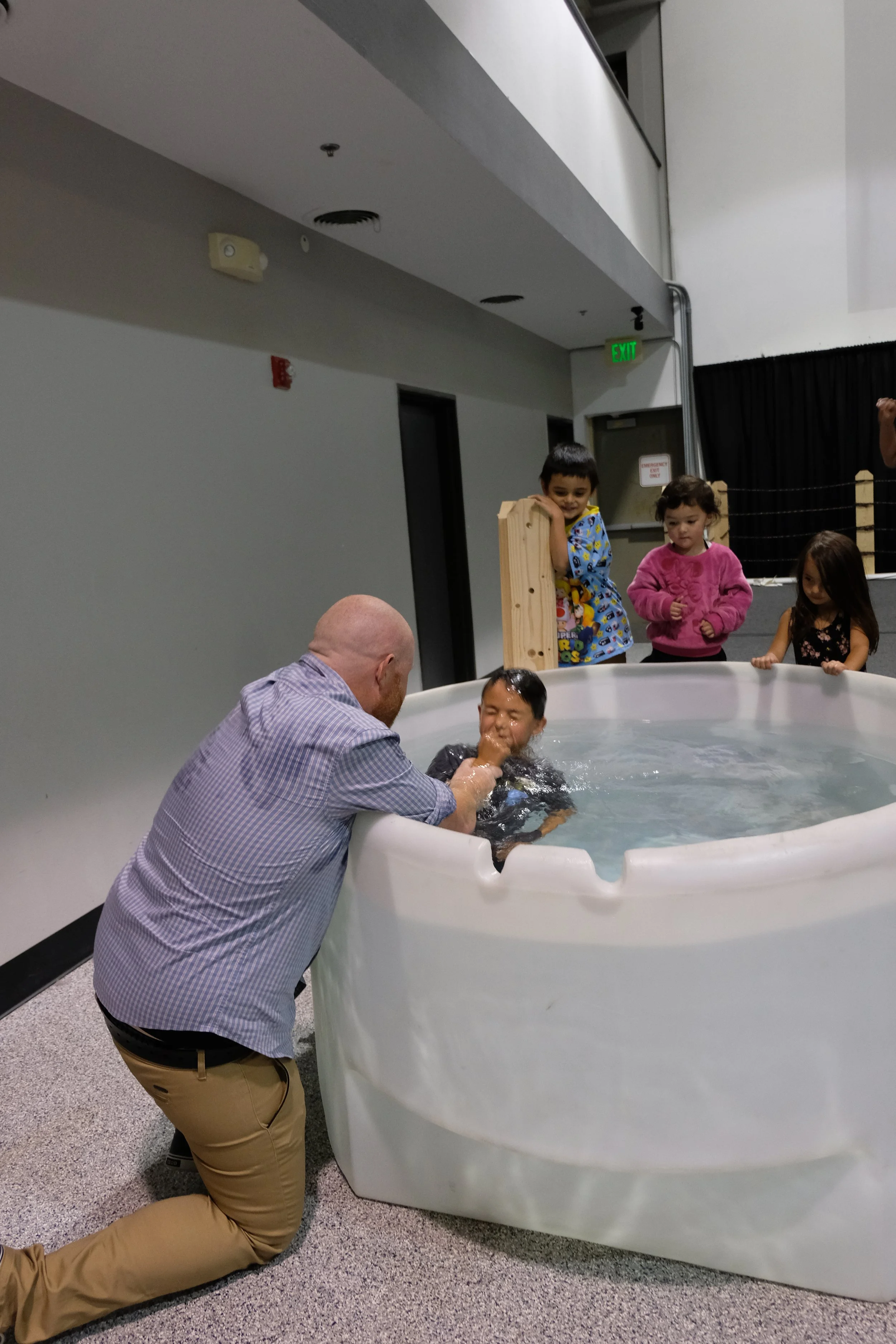 Young children watch as a man is baptized in a small white baptismal pool inside a room with gray walls and black curtain. The man is wet and being immersed by the man kneeling outside the pool.