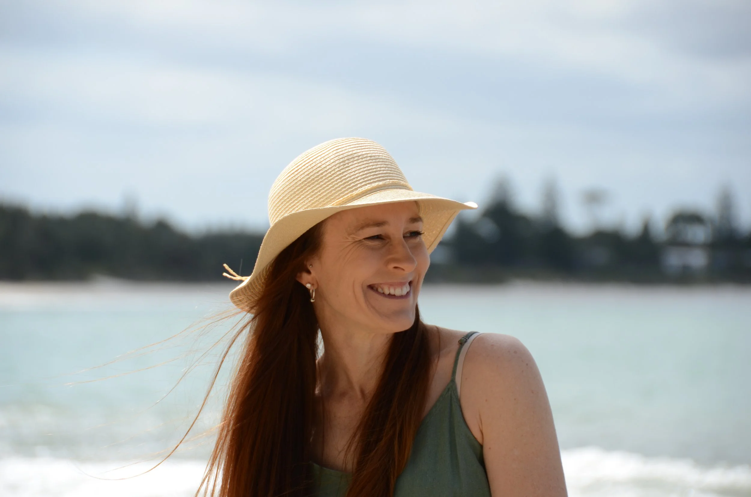 Woman with long hair wearing a straw hat and green tank top smiling at the beach with ocean and cloudy sky in the background.