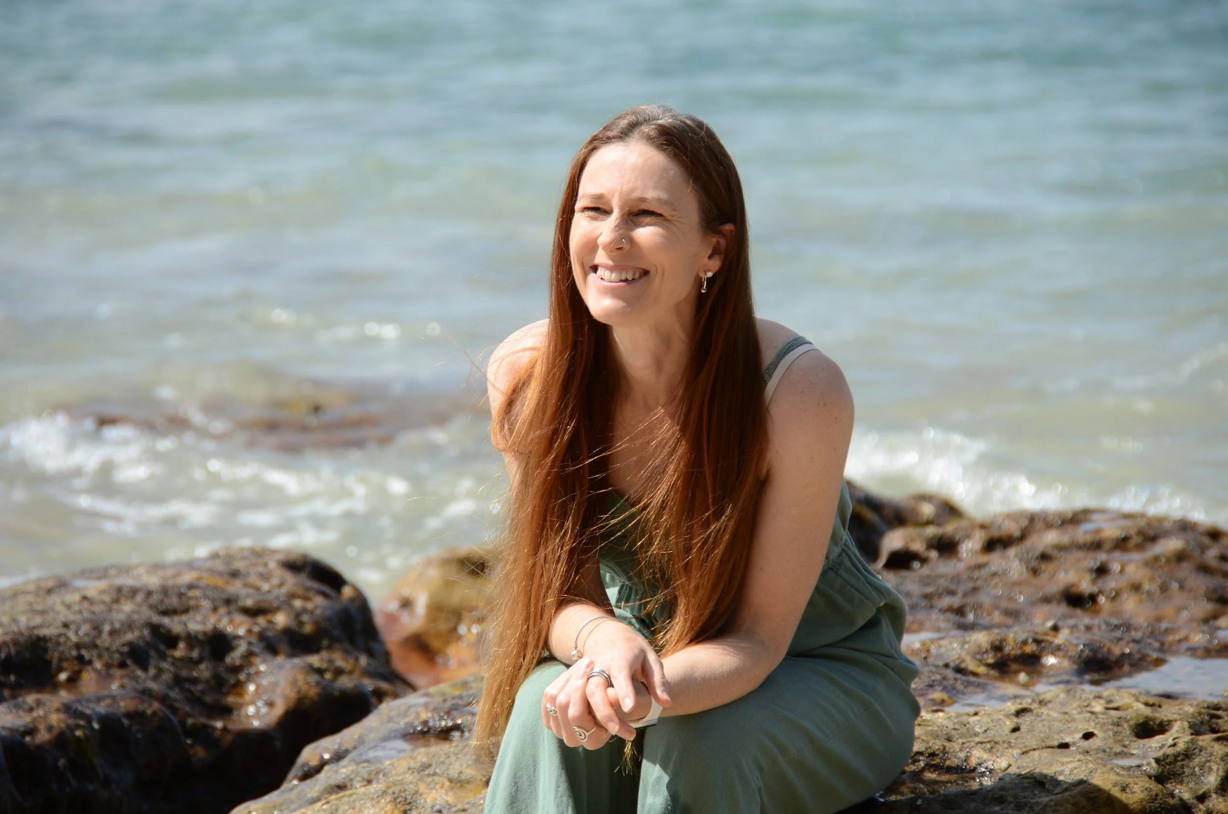 A woman with long red hair and a nose ring sitting on rocks by the water, smiling at the camera on a sunny day.