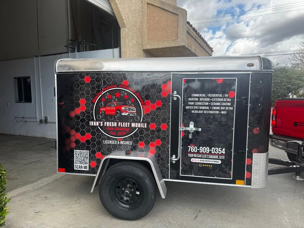 A black mobile detailing trailer with red and white graphics, including a red truck and honeycomb patterns, parked outside a building. The trailer displays contact information and services offered for auto detailing.