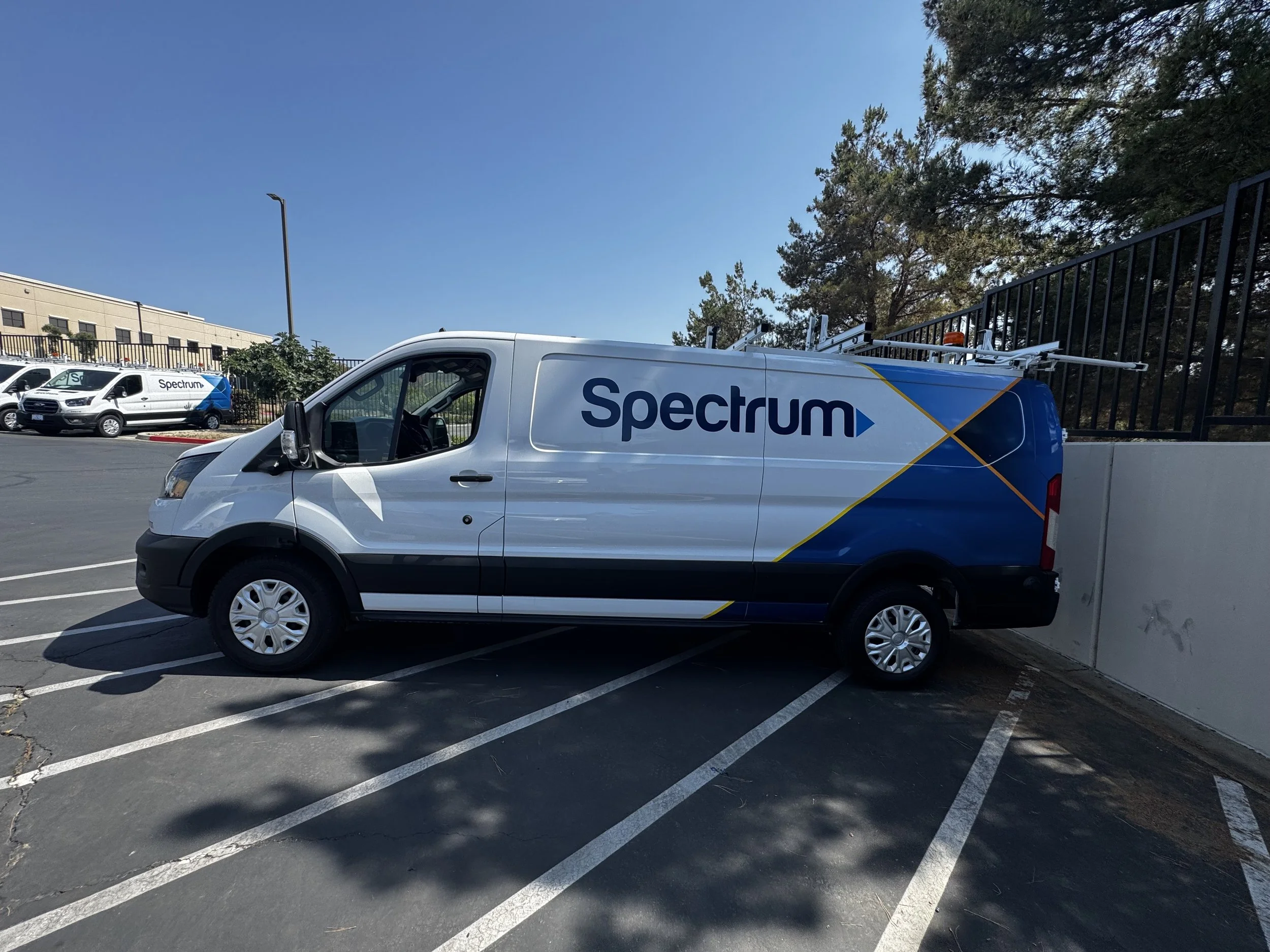 Spectrum branded service van parked in a parking lot near a concrete wall, with additional Spectrum vehicles in the background and a clear blue sky overhead.
