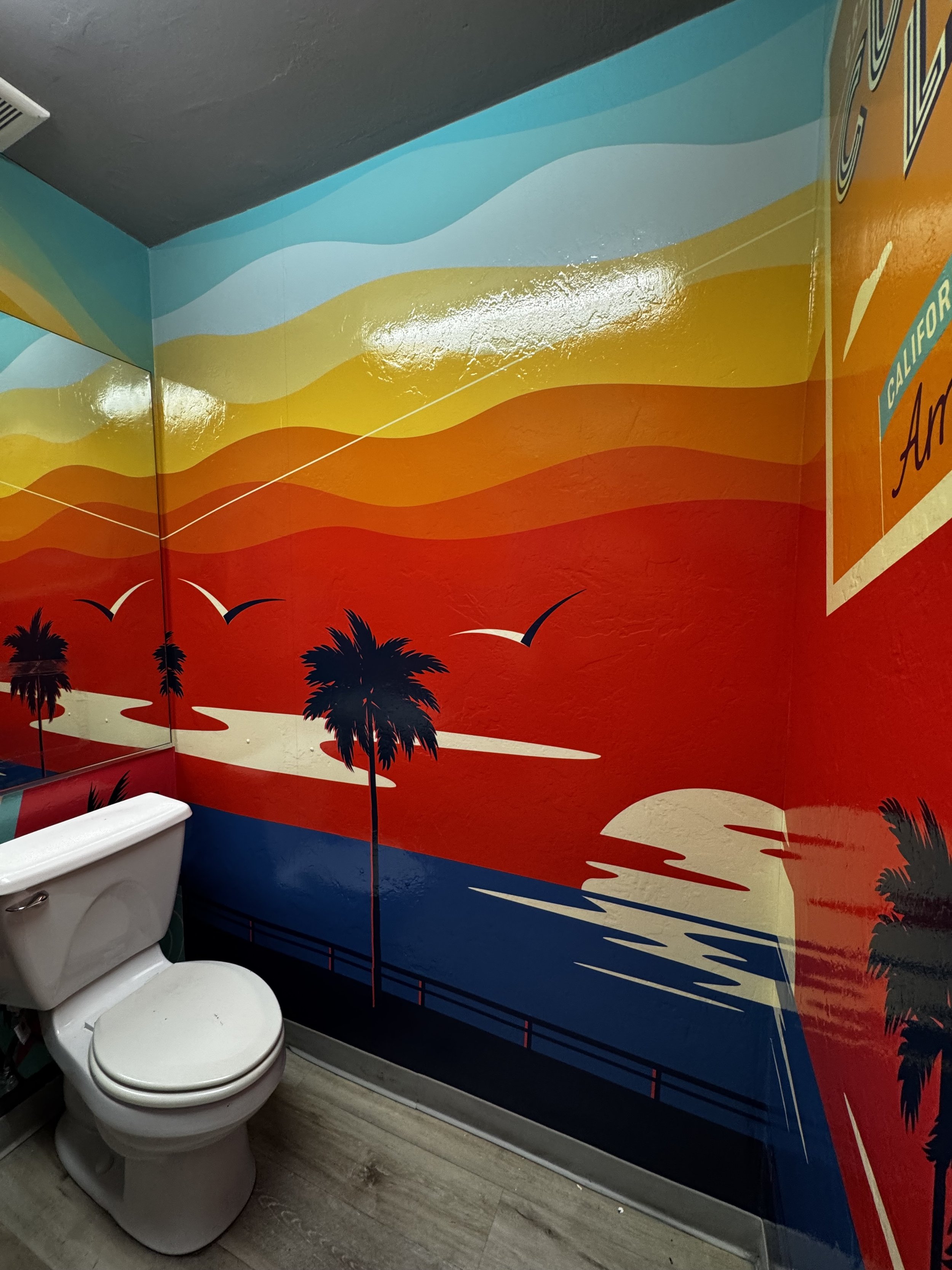 Colorful wall mural with sunset, palm trees, and seagulls, located in a bathroom next to a toilet.