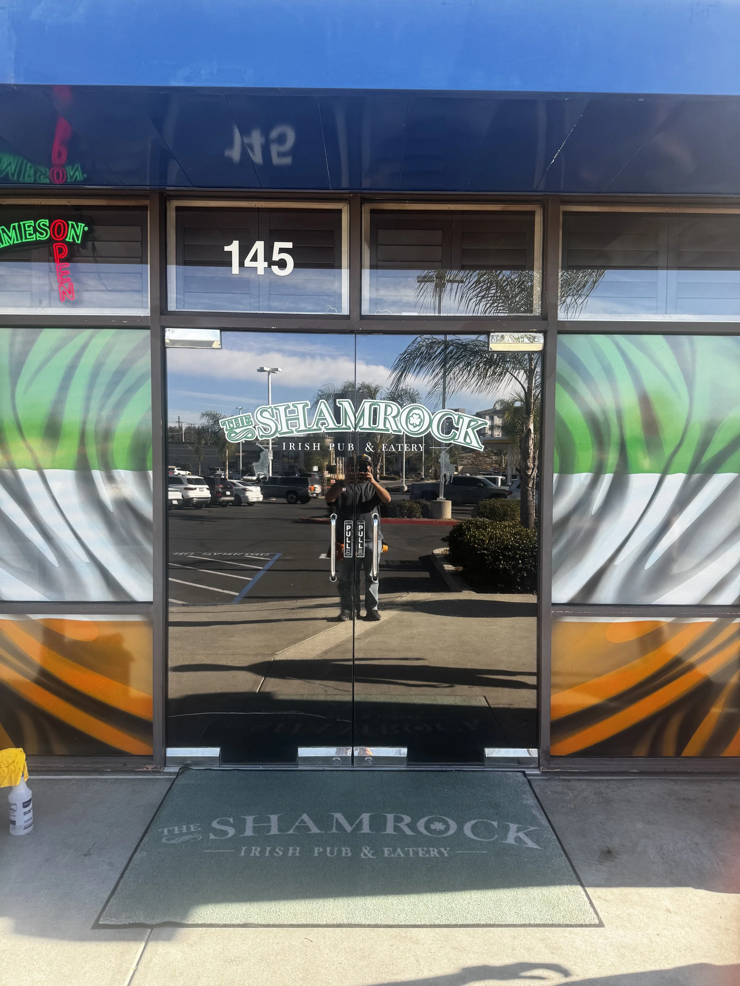 Front entrance of The Shamrock Irish Pub & Eatery with glass doors, a green and white logo, and a welcome mat. The reflection in the glass shows a parking lot, cars, palm trees, and a person taking the photo.