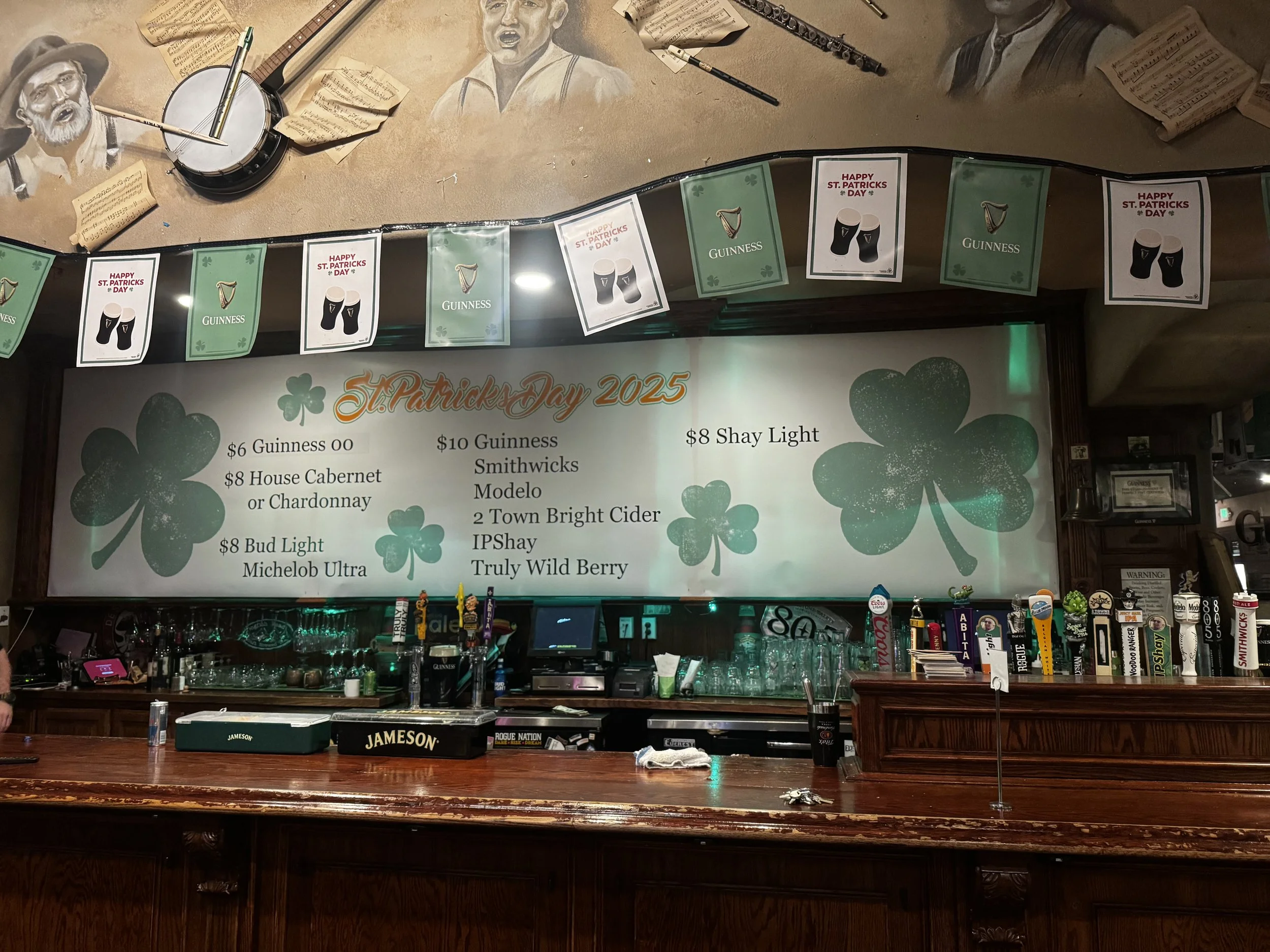A bar decorated for St. Patrick's Day 2025 with a menu listing various drinks and colorful shamrock graphics. The top features banners with beer and Guinness images and 'Happy St. Patrick's Day' messages.