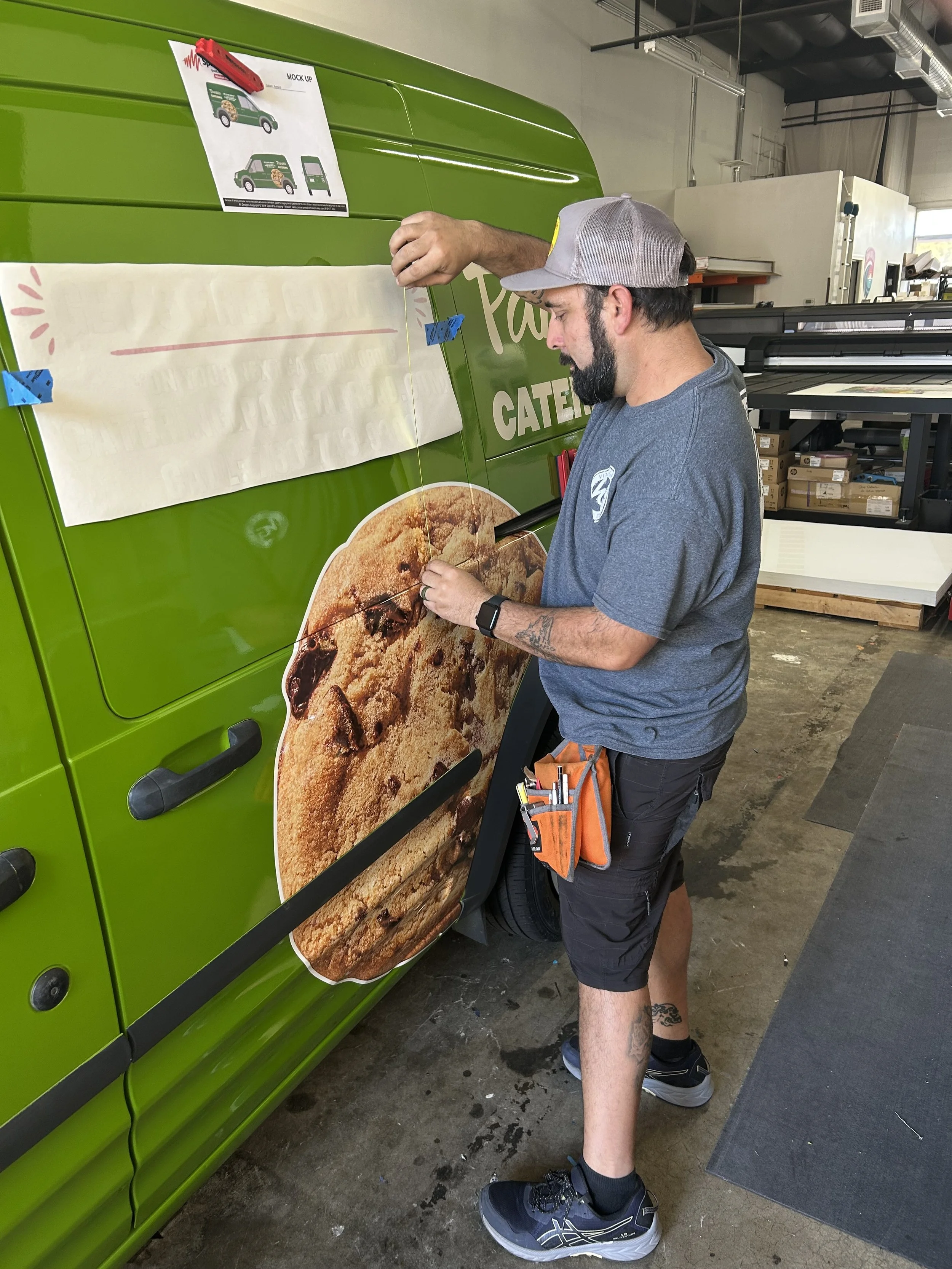 A man in a gray cap and a blue t-shirt is working on a green vehicle with a large cookie image on its side. He has tools attached to his pocket and is applying a decal or wrapping to the vehicle in an industrial workspace.