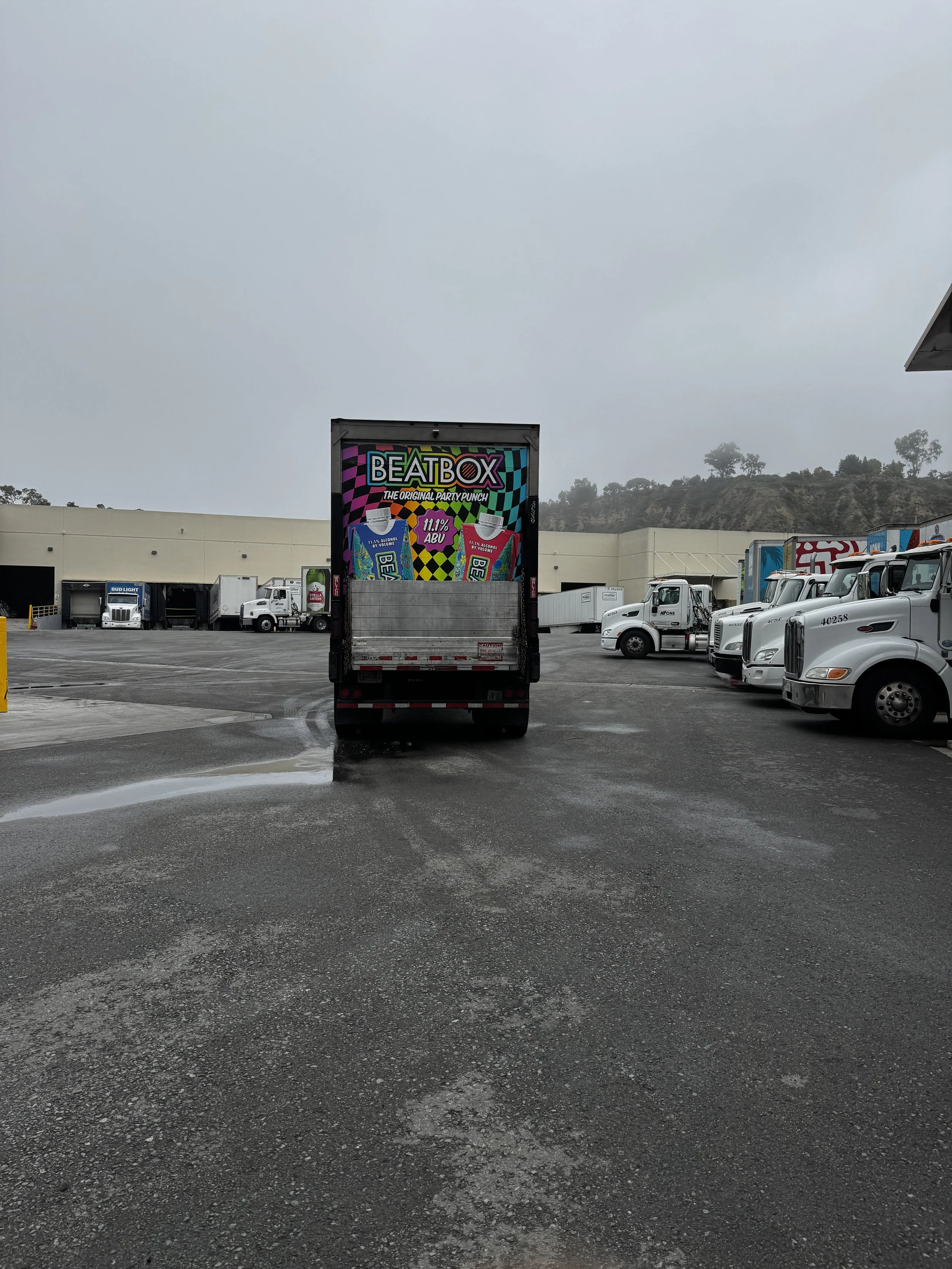 A delivery truck with an advertisement for BeatBox flavored beverage on its back, parked in a lot with other trucks and semi-trailers on a cloudy day.