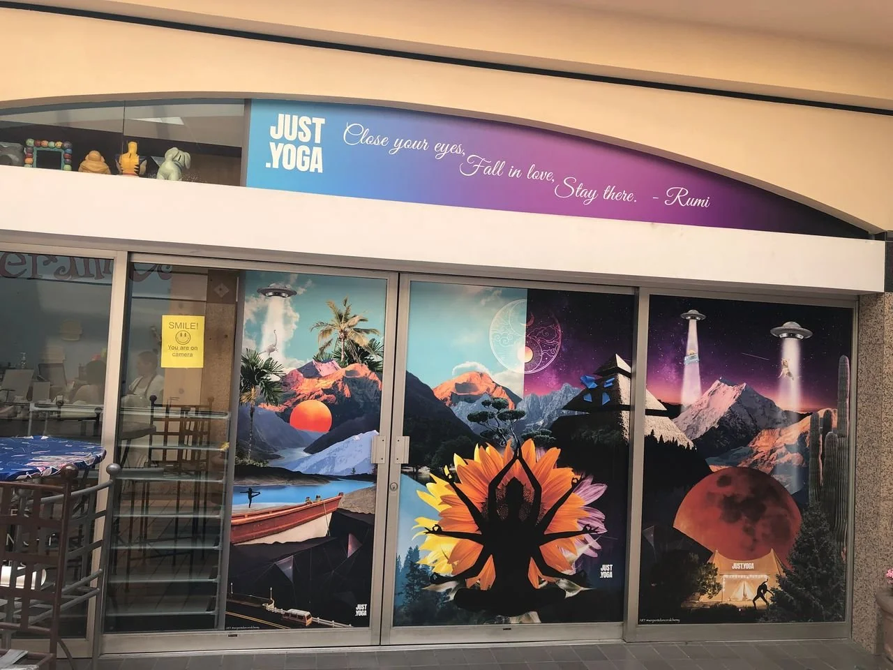 Colorful window mural depicting a mountainous landscape with a yoga silhouette in front of a large sunflower. The scene includes mountains, a sunset, a spaceship, UFOs, a full moon, and stars, creating a surreal, cosmic atmosphere.