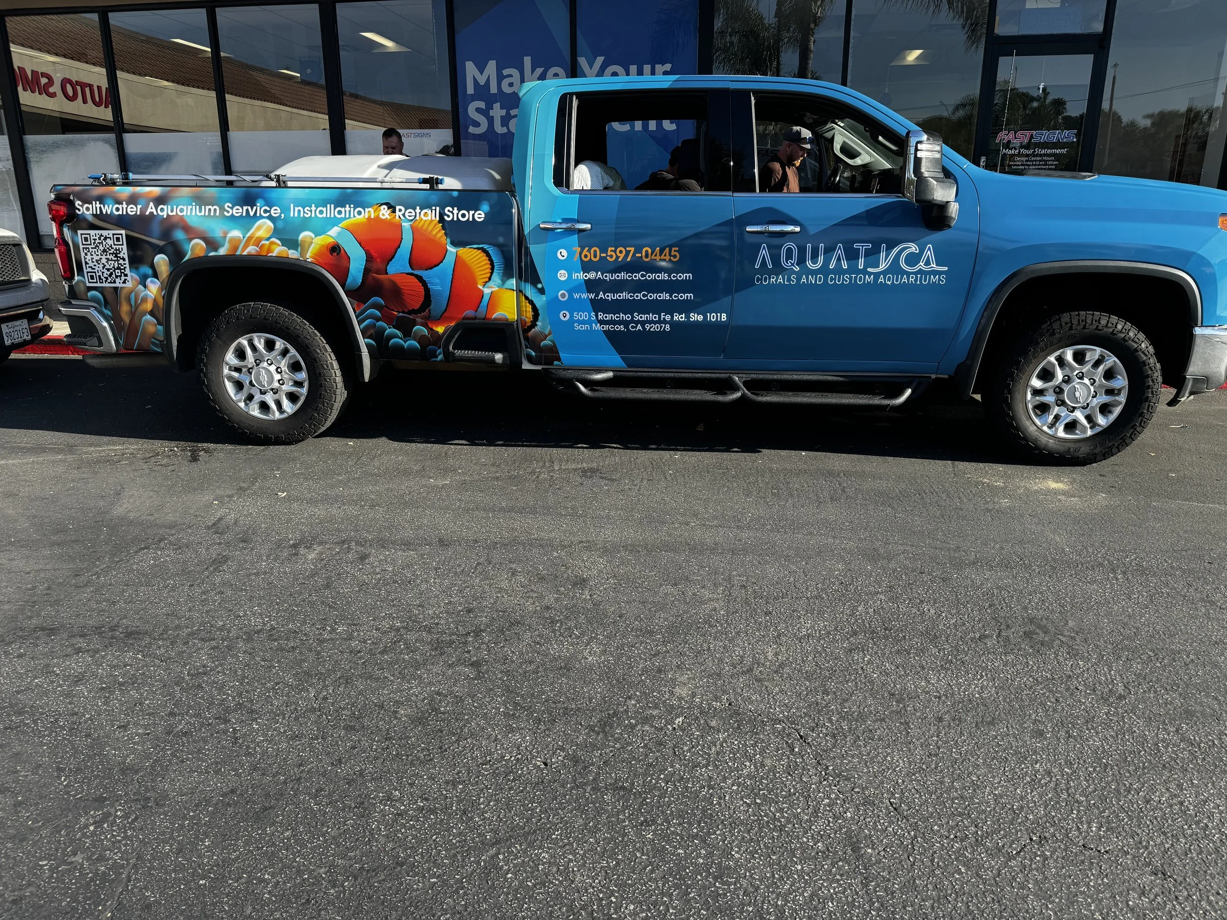 Blue pickup truck advertising Saltwater Aquarium Service, with colorful clownfish and coral reef graphics, parked outside a store with glass windows.