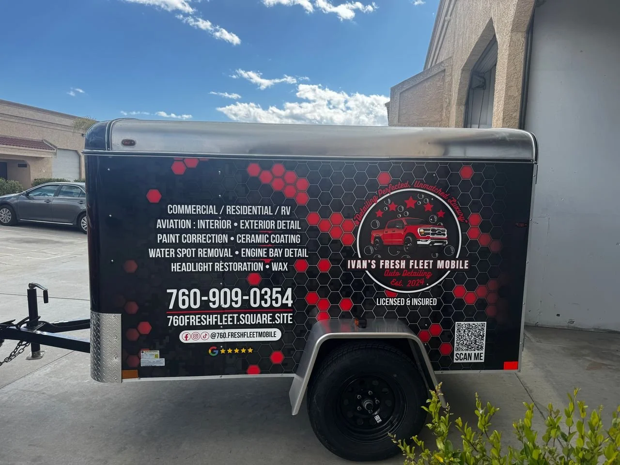 Black mobile auto detailing trailer with red and white text, hexagonal pattern design, and company logo featuring a red truck. Details include services offered, contact number, website, and social media icons. QR code and 