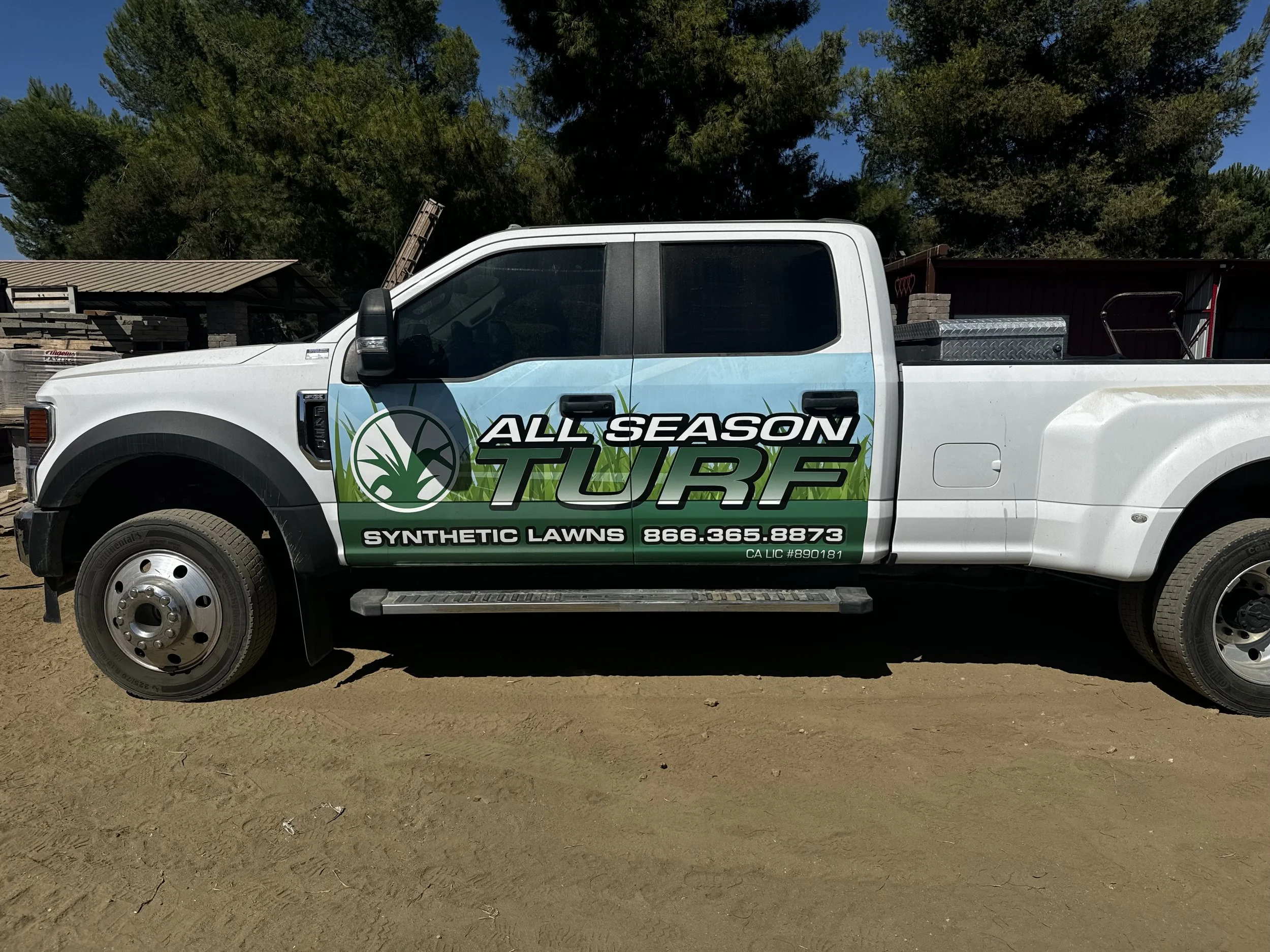 White pickup truck with a company logo and contact information on the side, parked on a dirt surface outdoors with trees and structures in the background.