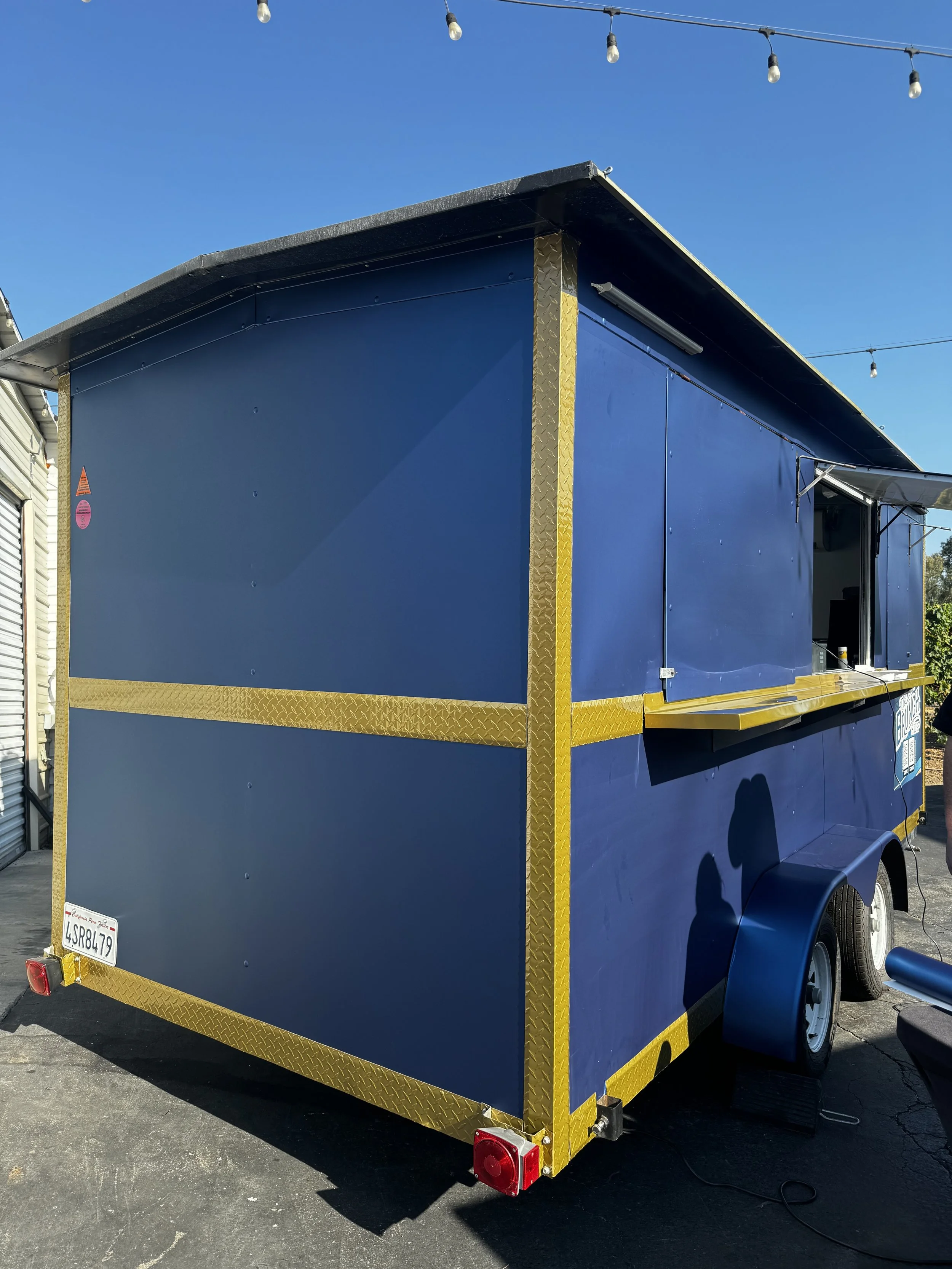 Blue food concession trailer with yellow trim and a serving window, parked outside under a string of lights on a sunny day.