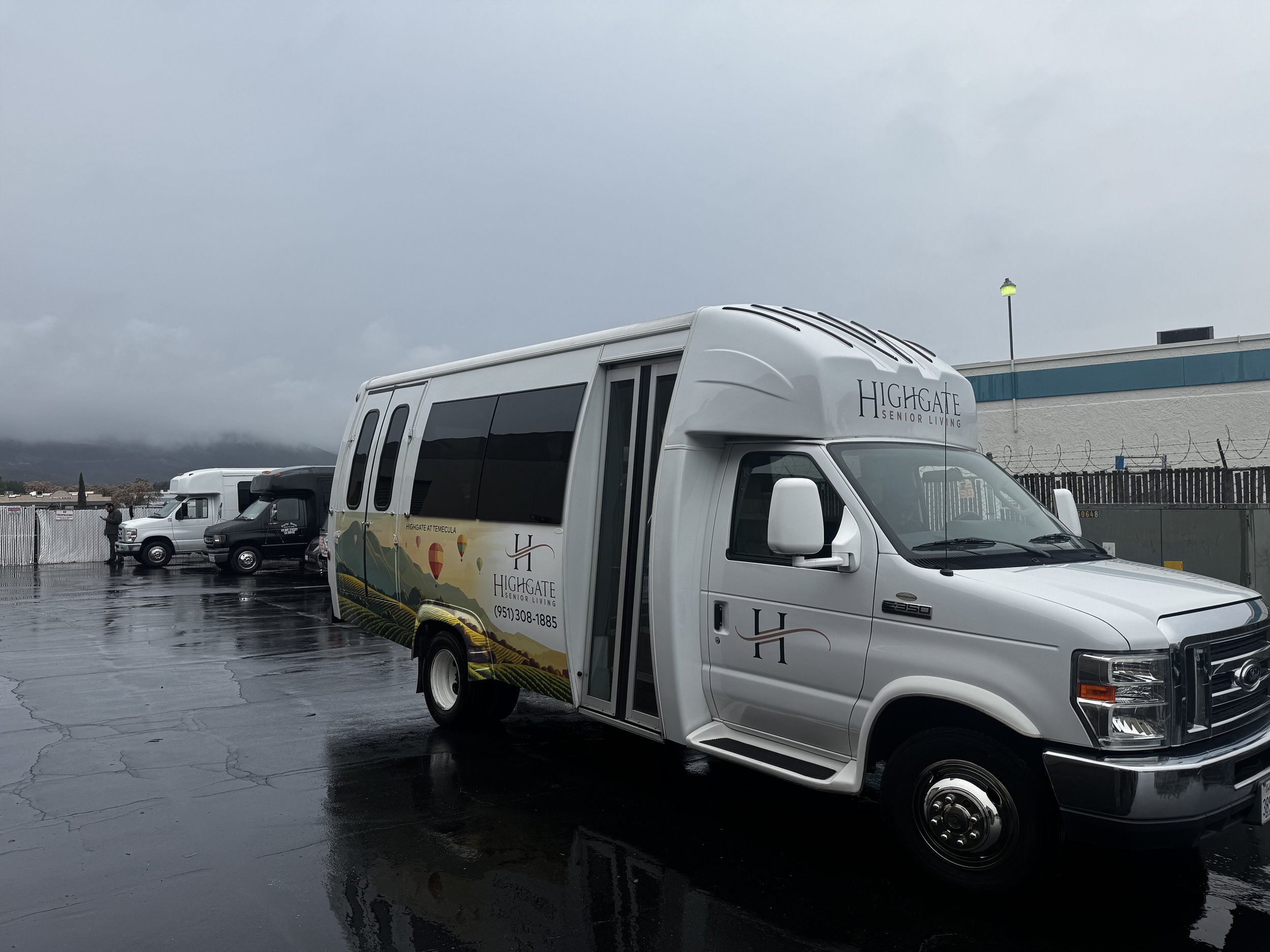 A white van with branding for Highgate Senior Living parked on a wet asphalt surface, with other similar vans in the background under cloudy skies.