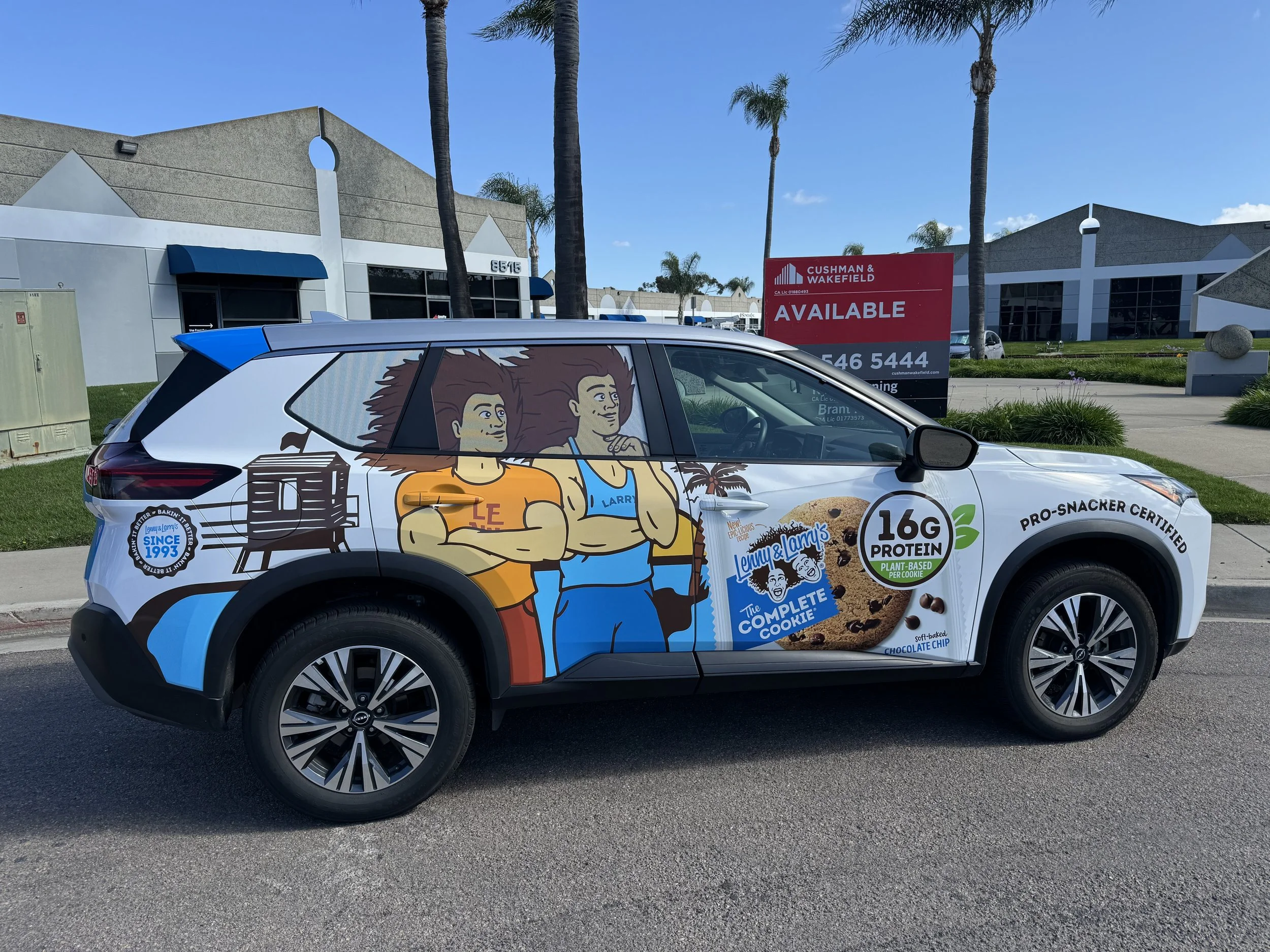 Custom car wrap featuring animated characters of a boy and a girl with crossed arms, advertising Lenny & Larry's chocolate chip cookies with nutritional claims like 16g protein and plant-based ingredients, parked on a street in front of commercial bu