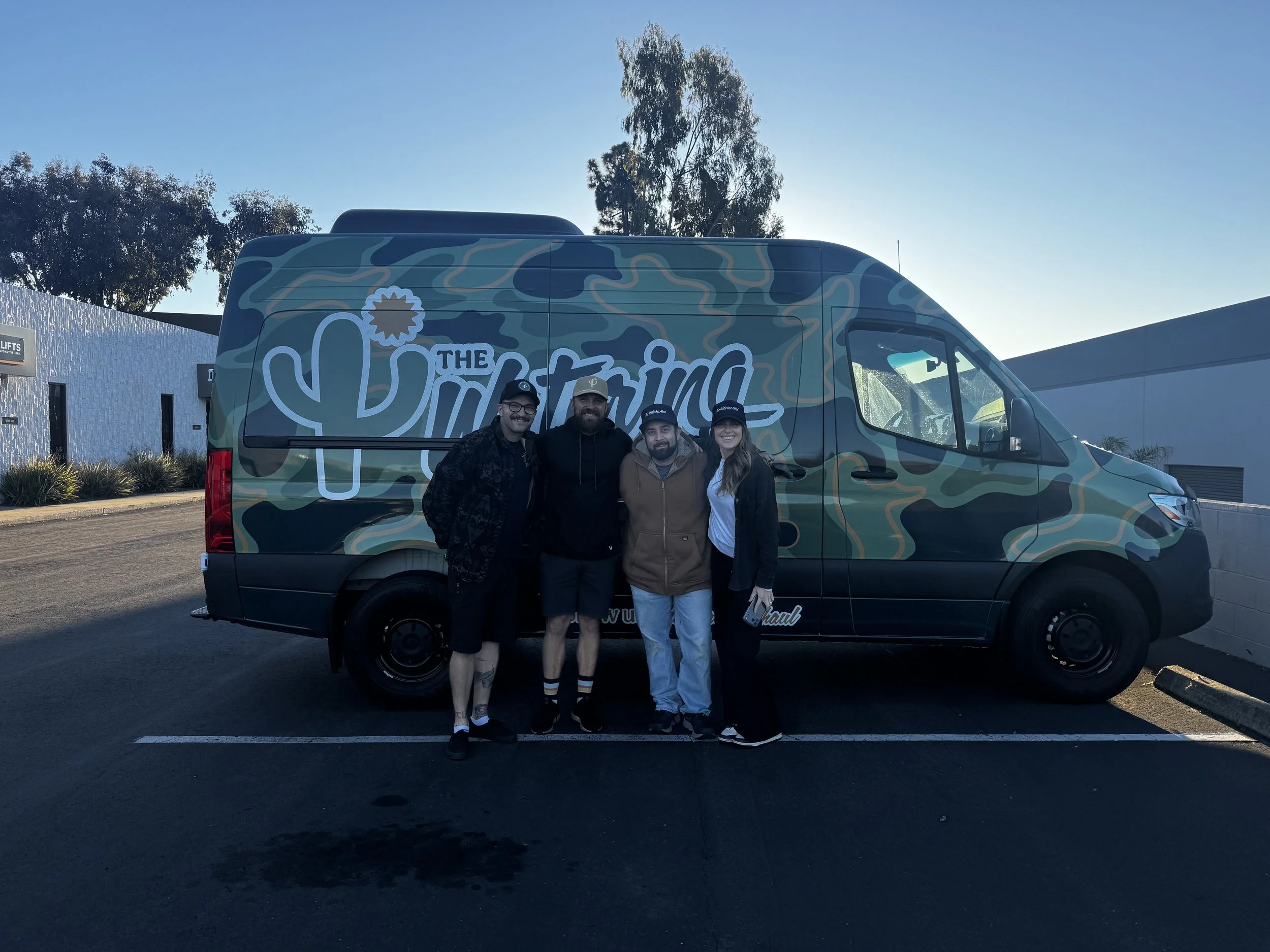 Four people standing in front of a camouflage van with Arizona-themed graphics and the words 'The Arizona' on the side, in a parking lot during daytime.
