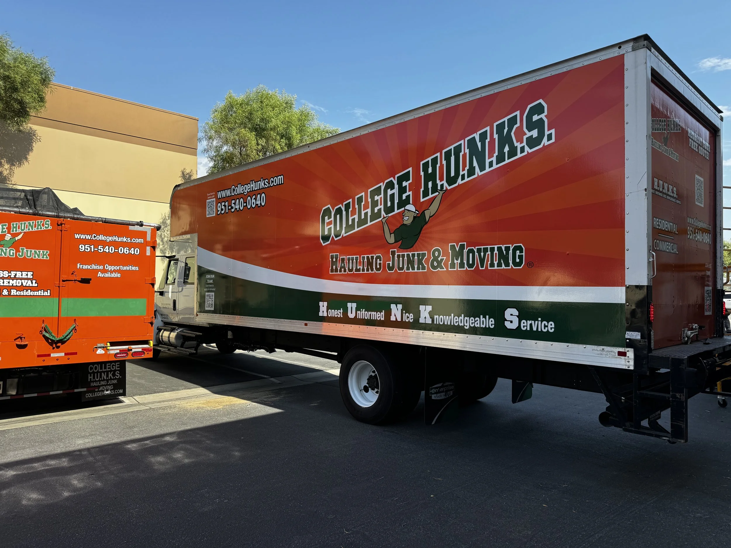 Two trucks parked in a lot with branding for College Hunks Hauling Junk and Moving. The larger truck is mainly red with white and green accents, featuring the company logo and contact information. The smaller truck is orange with green and white acce