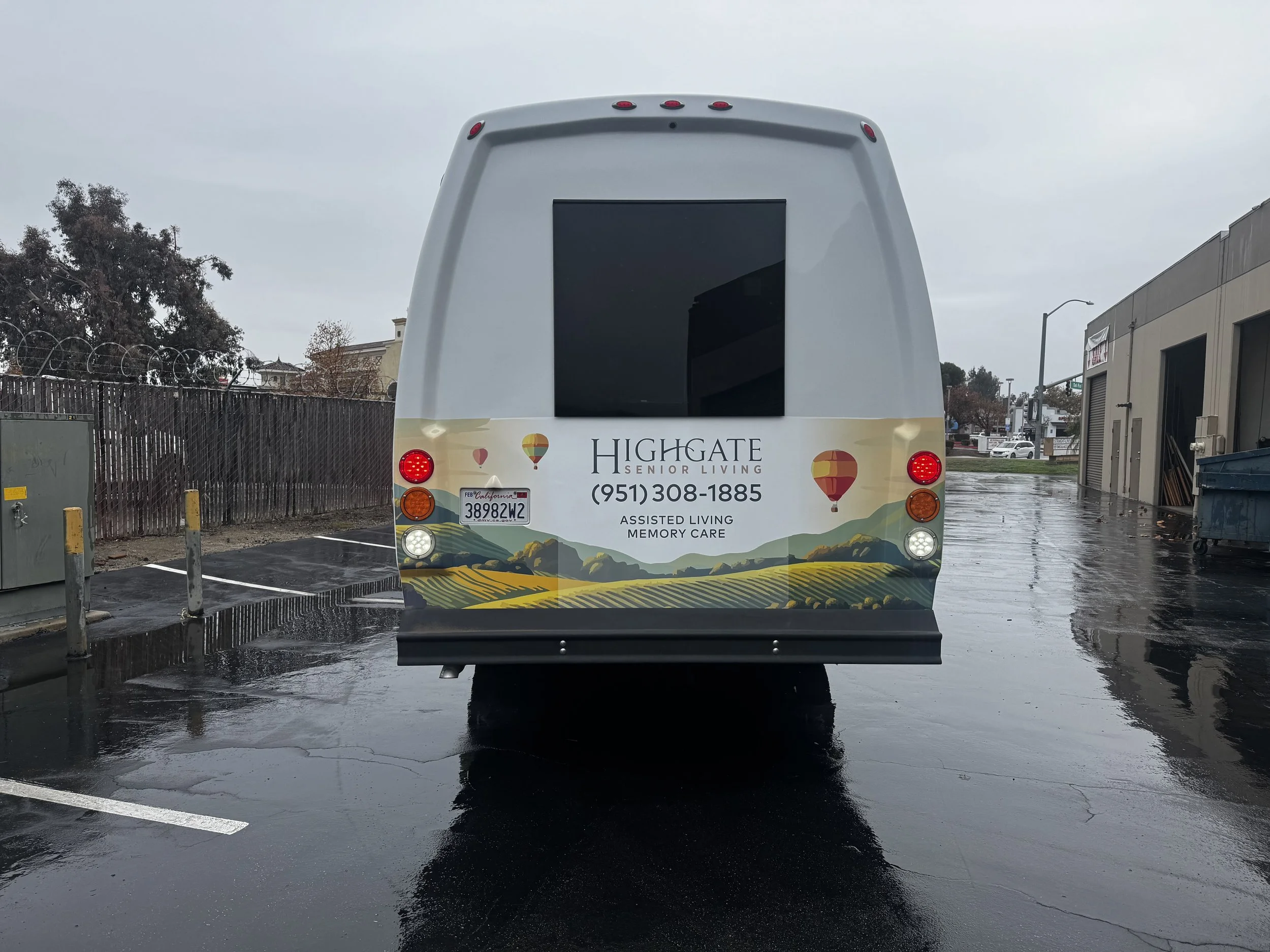 Rear view of a white senior living bus with a landscape illustration and hot air balloons, parked on wet pavement on a rainy day.