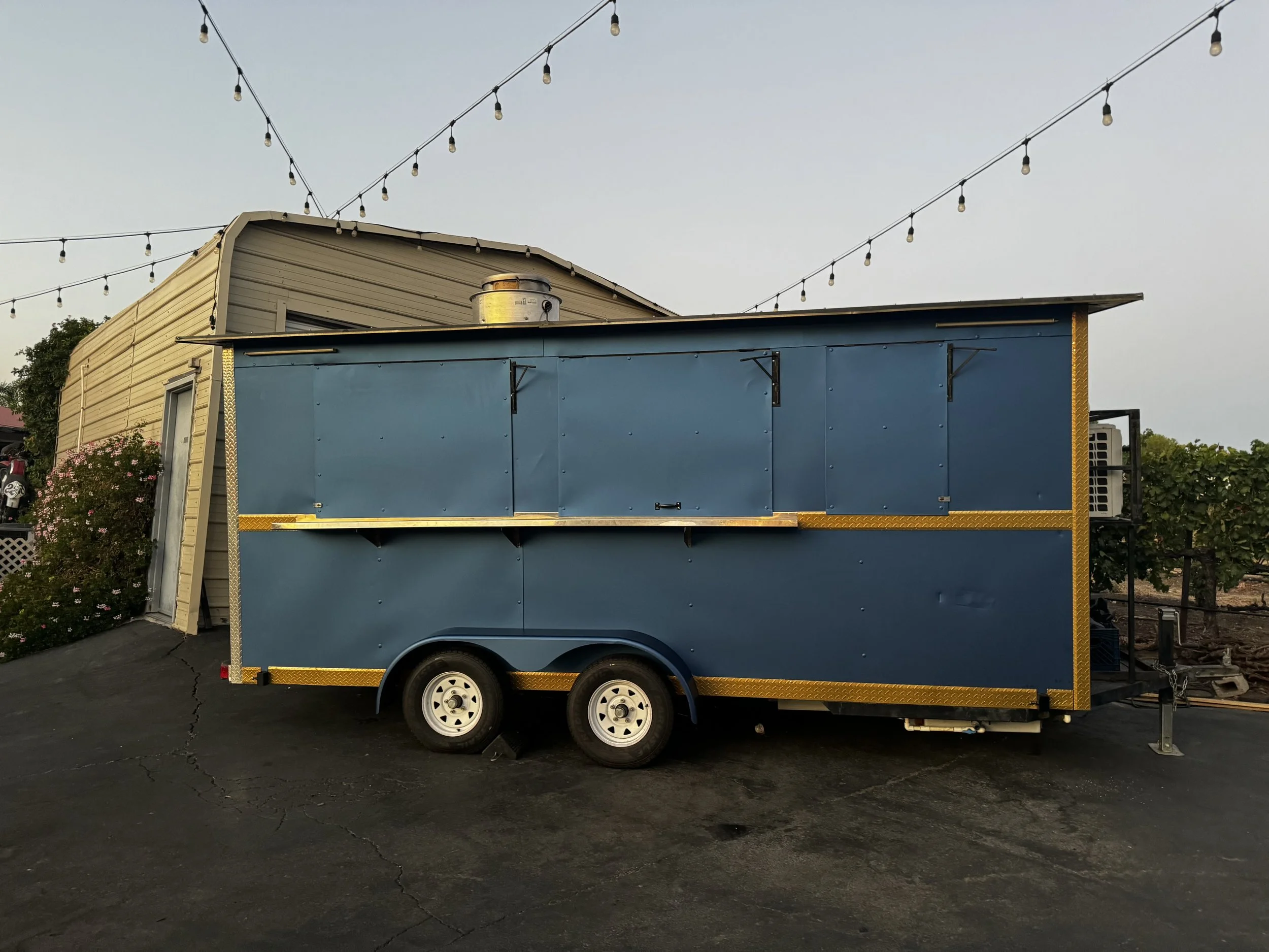 Blue food trailer with yellow trim parked on asphalt, with a beige building and string lights overhead.