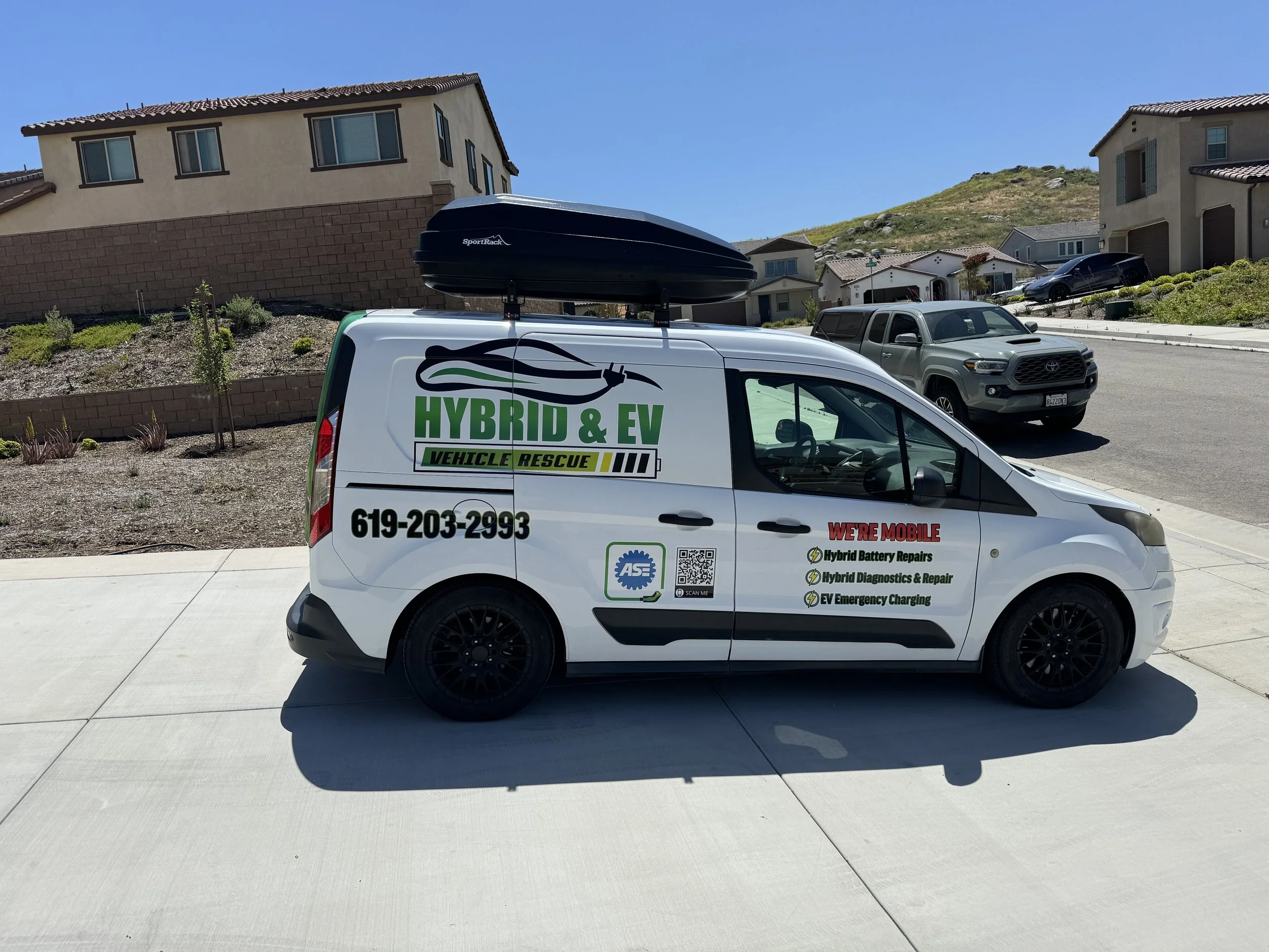 A white van with green and black company logos and text parked on a driveway in a suburban neighborhood. The van has a rooftop cargo box and contact information on its side, advertising hybrid and electric vehicle services.