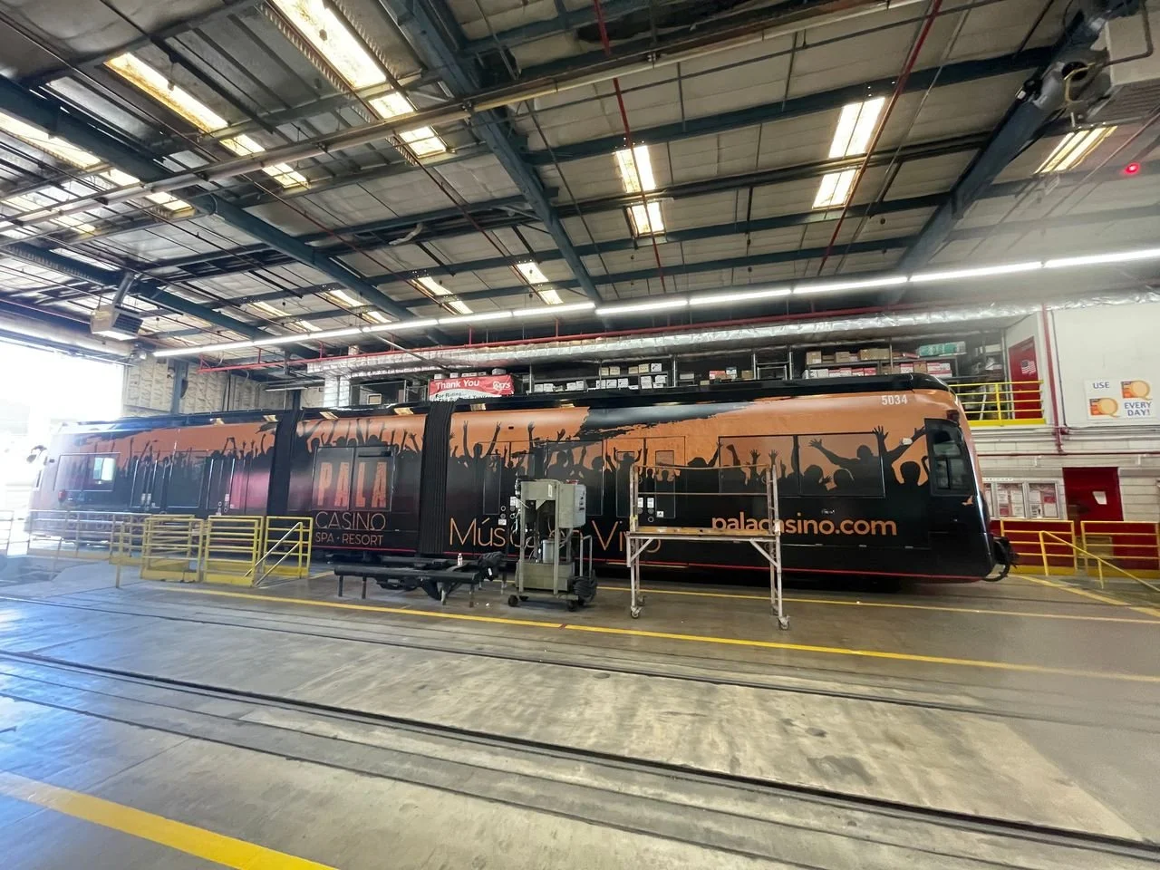 Inside a large industrial space with a bus or tram decorated with promotional artwork for Palms Casino Resort, featuring silhouettes of cheering people and the website palmcasino.com.