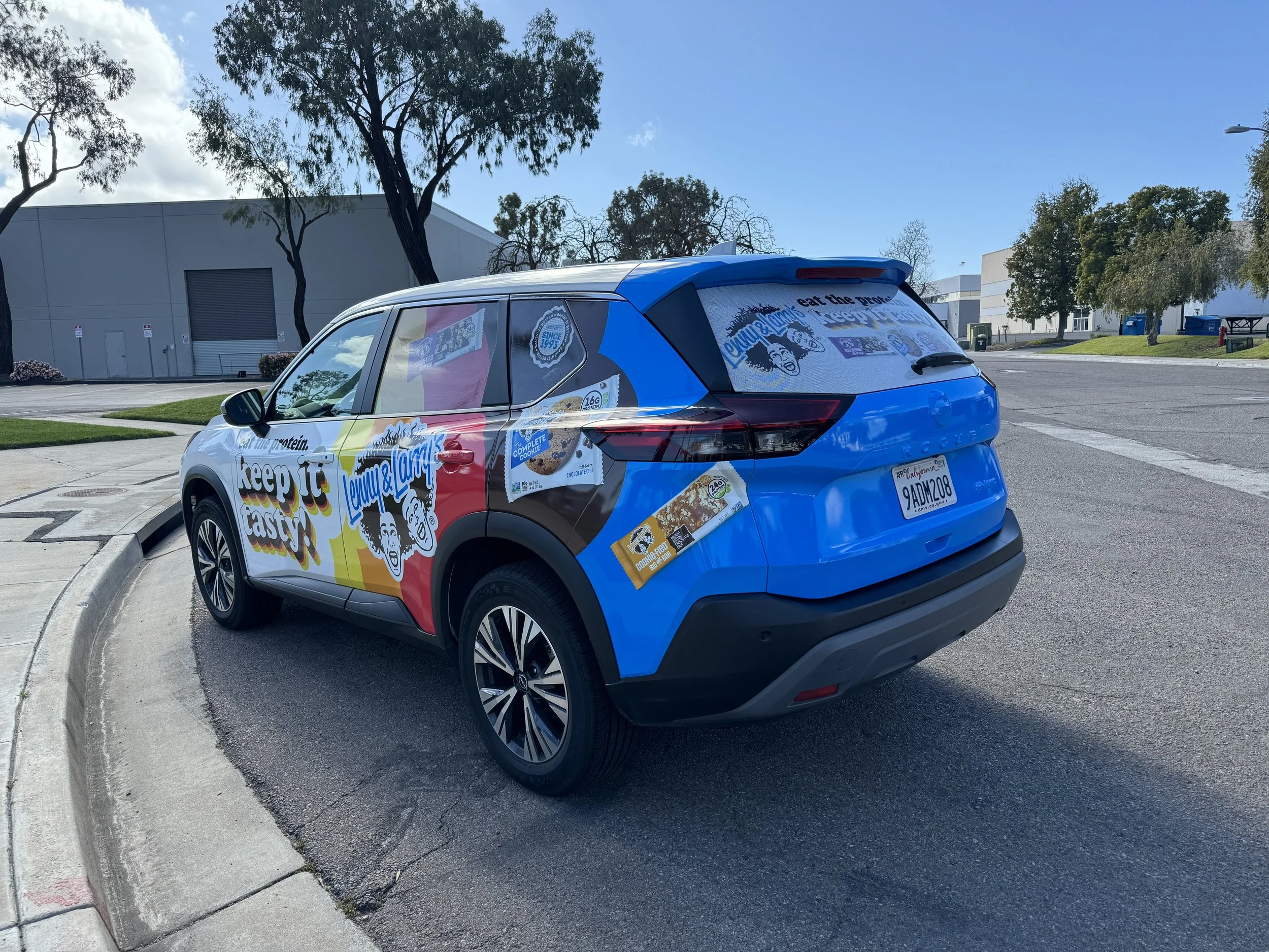 Blue car with colorful advertising decals for Layne & Lamy, promoting protein snacks on the sides and back, parked on the street near a sidewalk and trees.