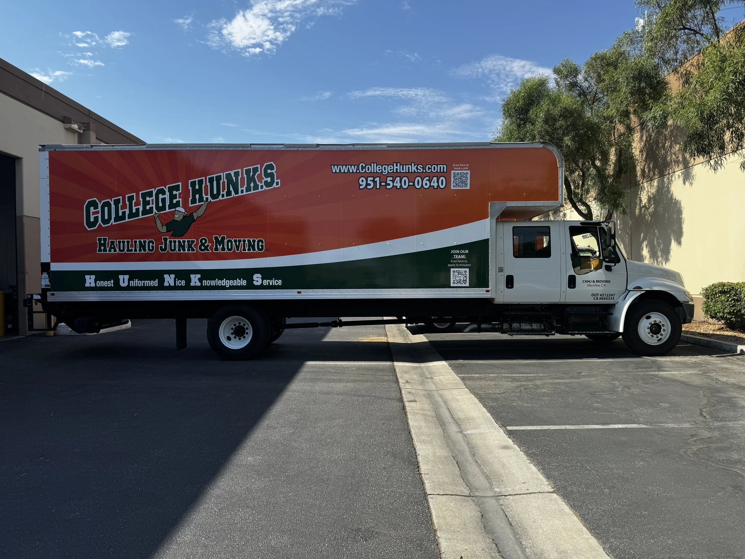 A large moving truck parked in a parking lot near a building, with trees and a blue sky in the background. The truck has branding for College Hunks Hauling Junk & Moving, including their website, phone number, and slogan.