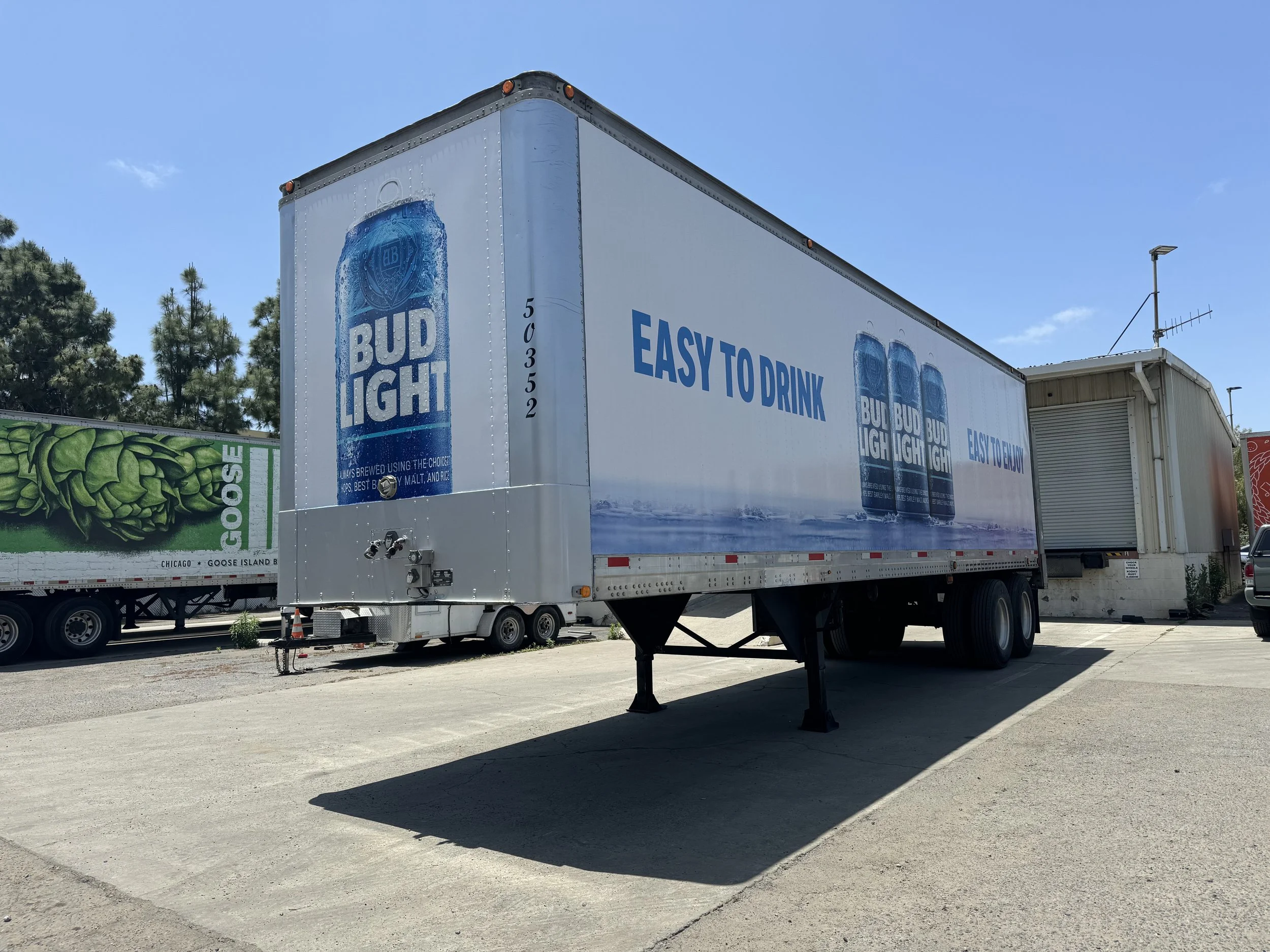 A large white truck trailer with an advertisement for Bud Light beer, displaying the slogan 'EASY TO DRINK' and an image of three cans of Bud Light on a water background.