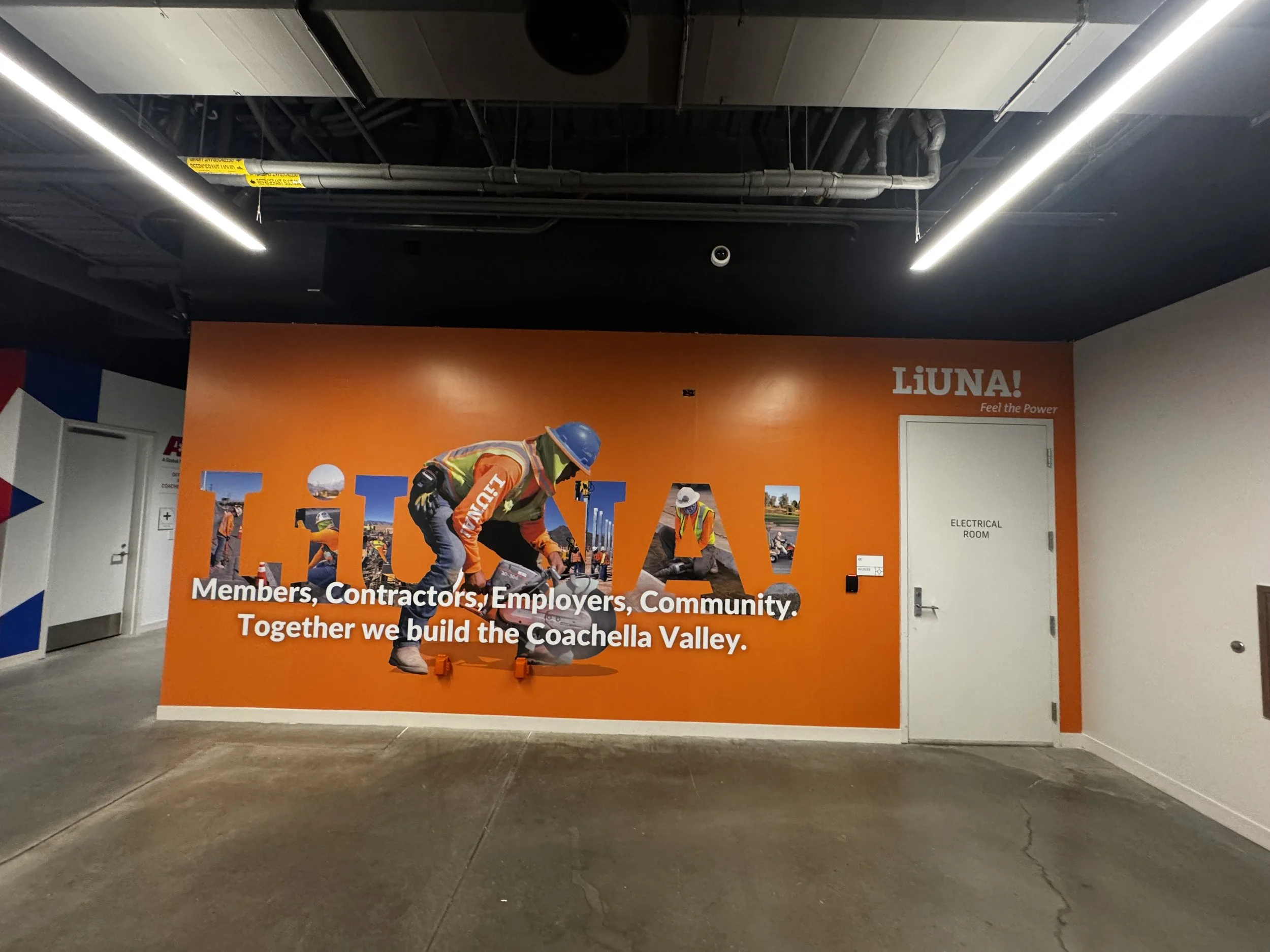 An orange wall with the word 'LiUNA!' and slogan 'Feel the Power' in the upper right corner. The wall features a mural of construction workers in safety vests and helmets, with a quote 'Members, Contractors, Employers, Community. Together we build th