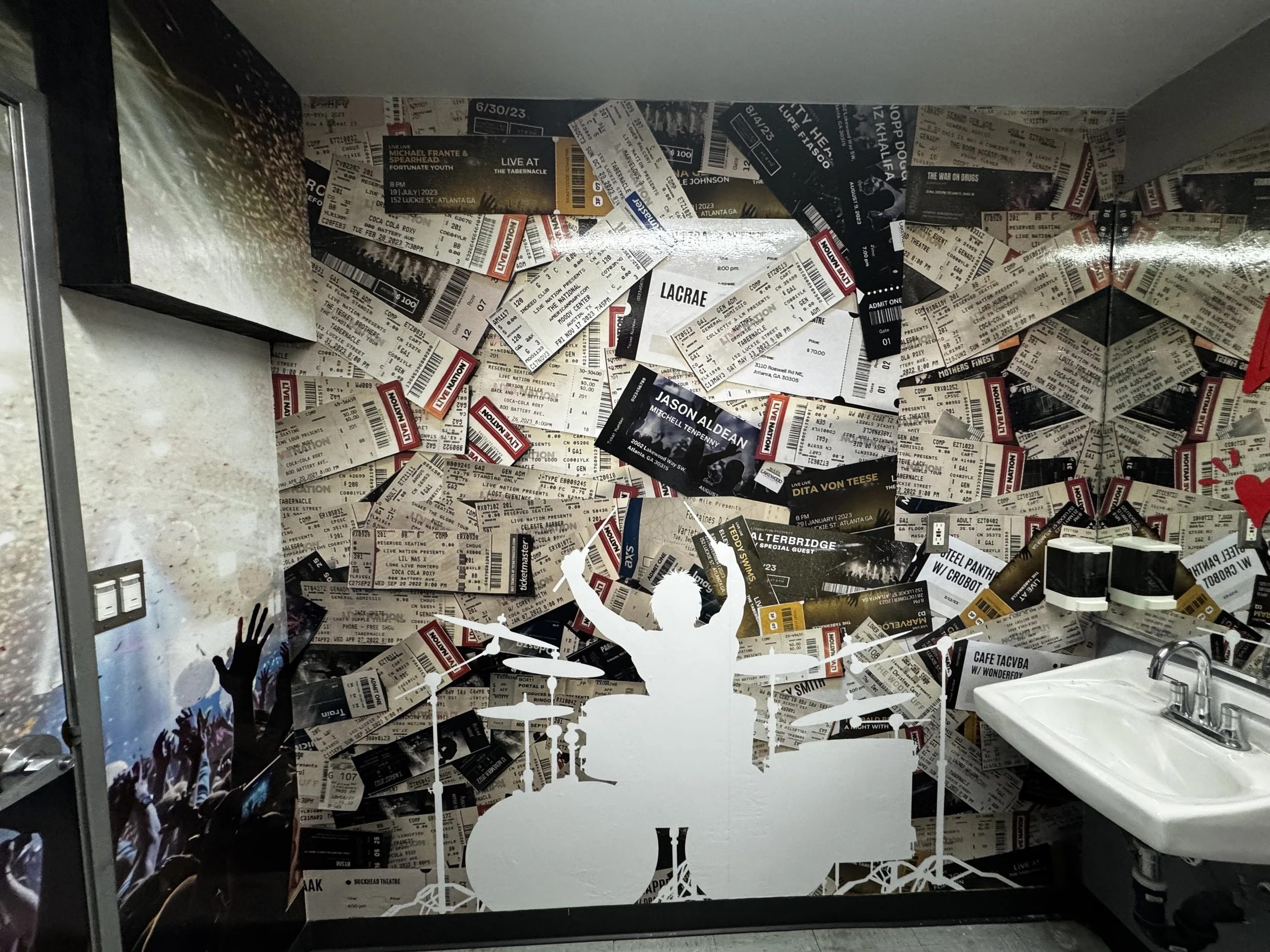 A wall decorated with a collage of concert tickets, tickets mostly overlapping and creating an abstract background, with a silhouette of a drummer playing drums in the foreground, and a crowd with raised hands also depicted in front of the wall.