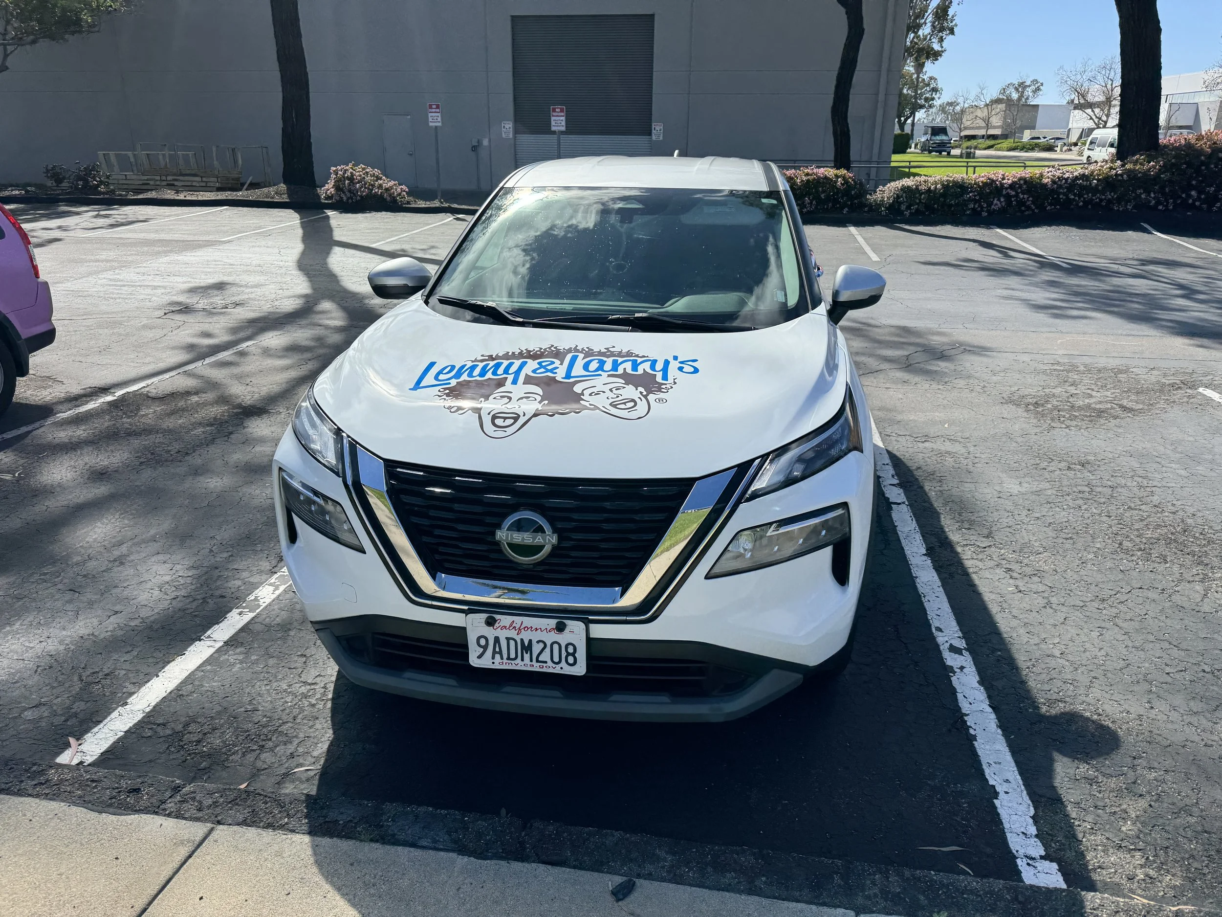 White Nissan SUV with a decal on the hood featuring the logo of Lenny & Larry's, a cookie company, in a parking lot.
