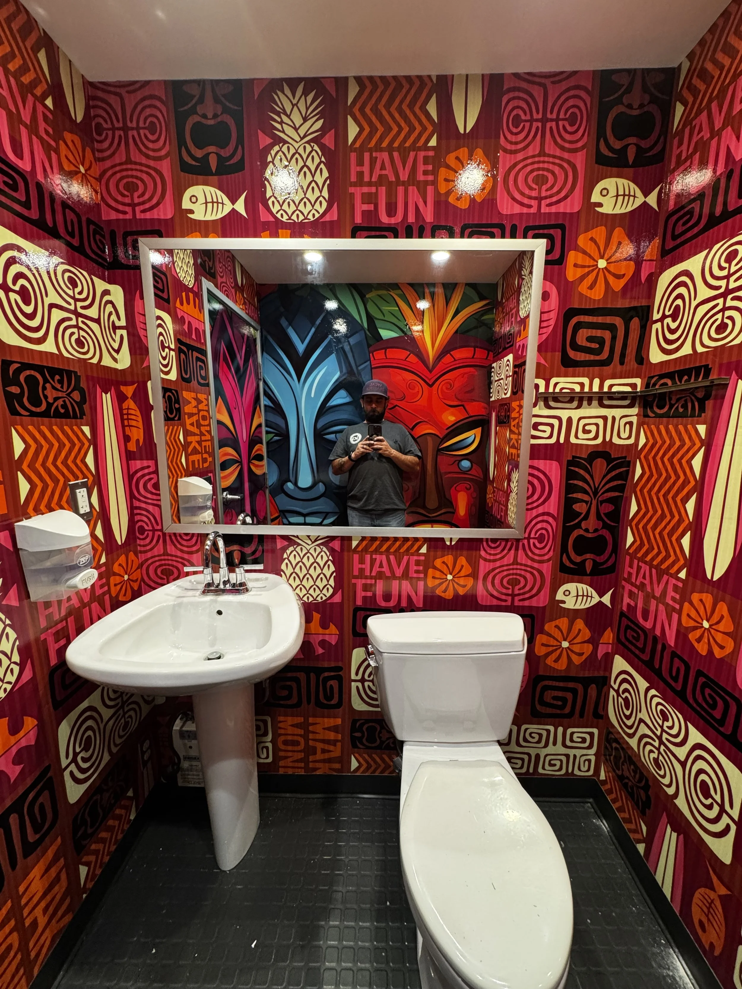 Bathroom with colorful mural wallpaper featuring digital art of masks, flowers, fish, and words like 'Have Fun' and 'Way Money.' White sink and toilet, black rubber mat on the floor, and a man taking a selfie in the mirror.